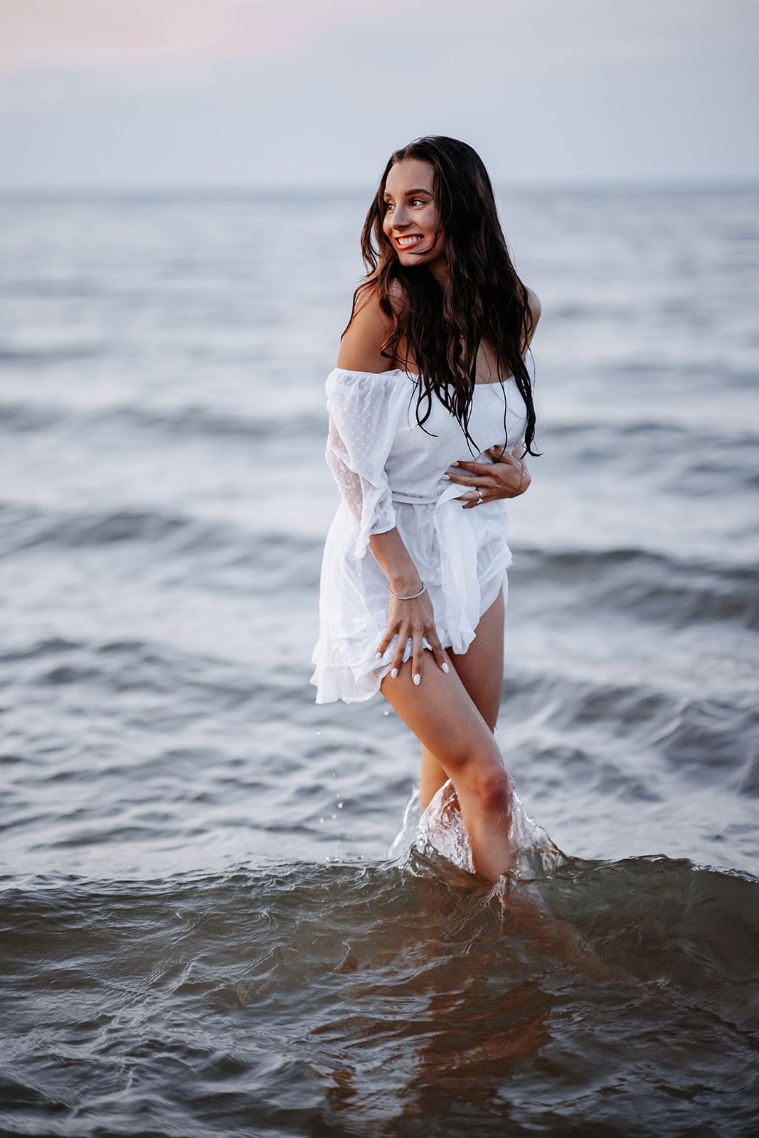 Smiling brunette woman in white off-shoulder dress standing in ocean waves at dusk.