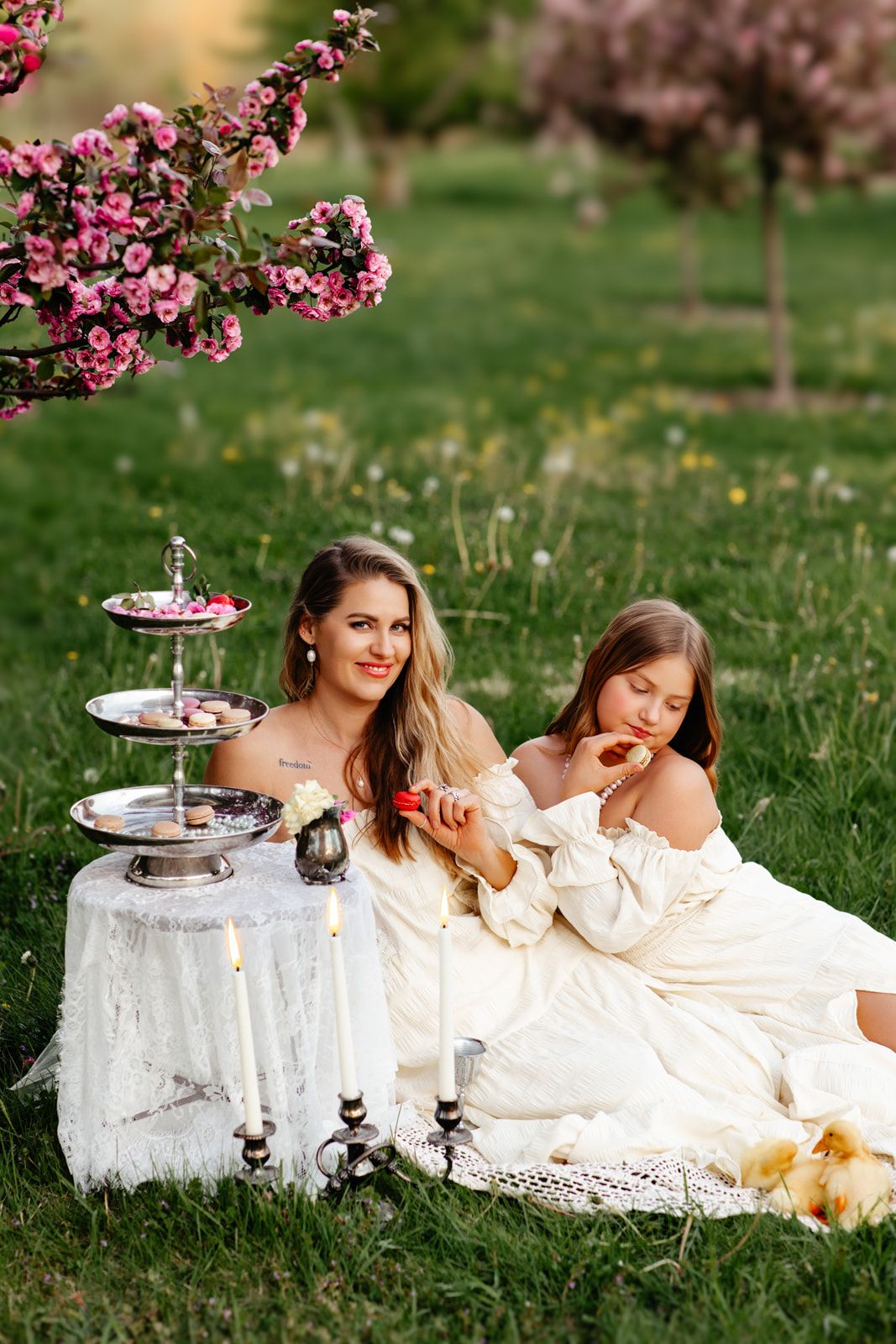 Mother and daughter in white dresses enjoying a picnic under blooming pink cherry trees on green grass.