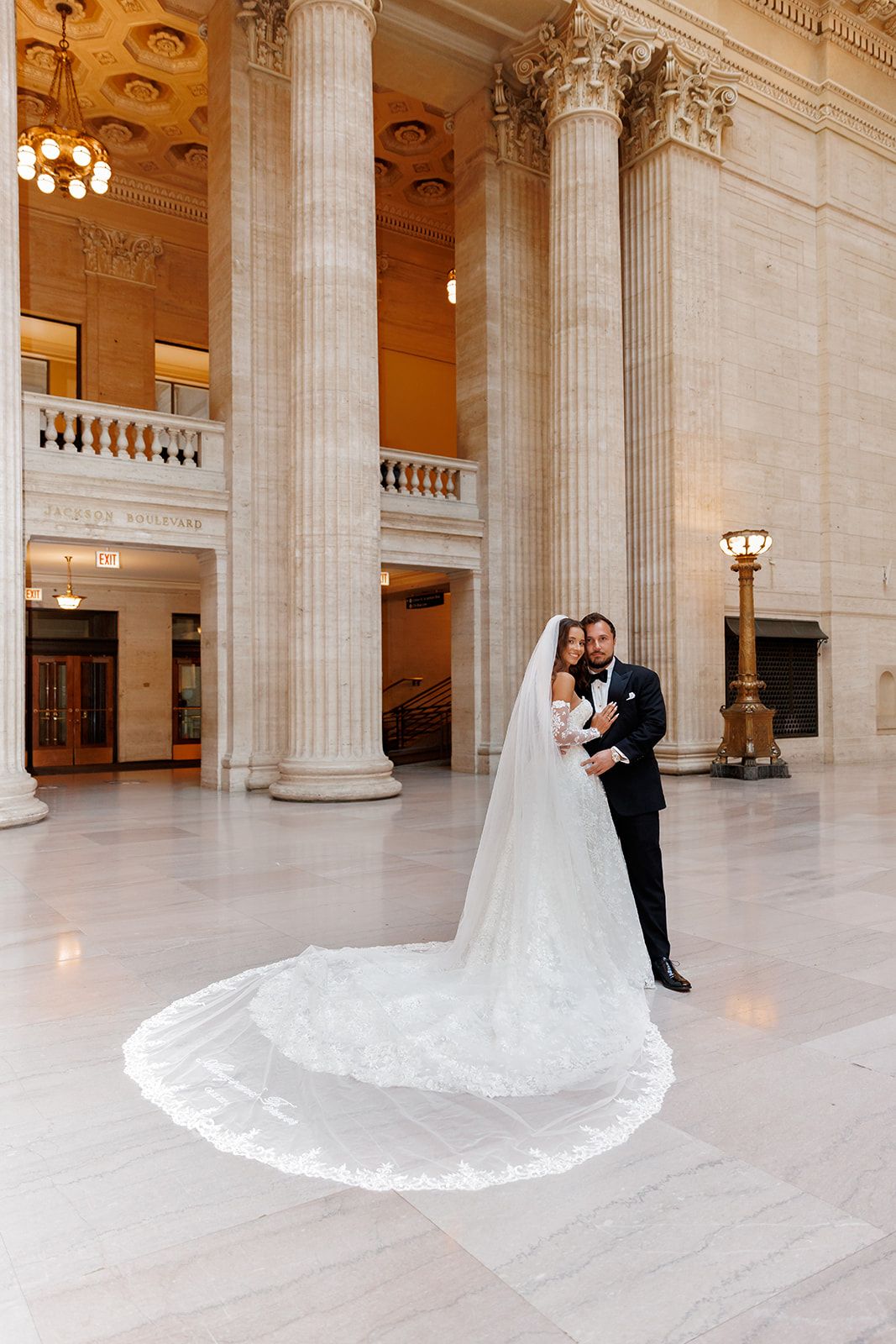 Bride in a long lace veil embraced by groom in tuxedo in a grand marble hall with tall columns.