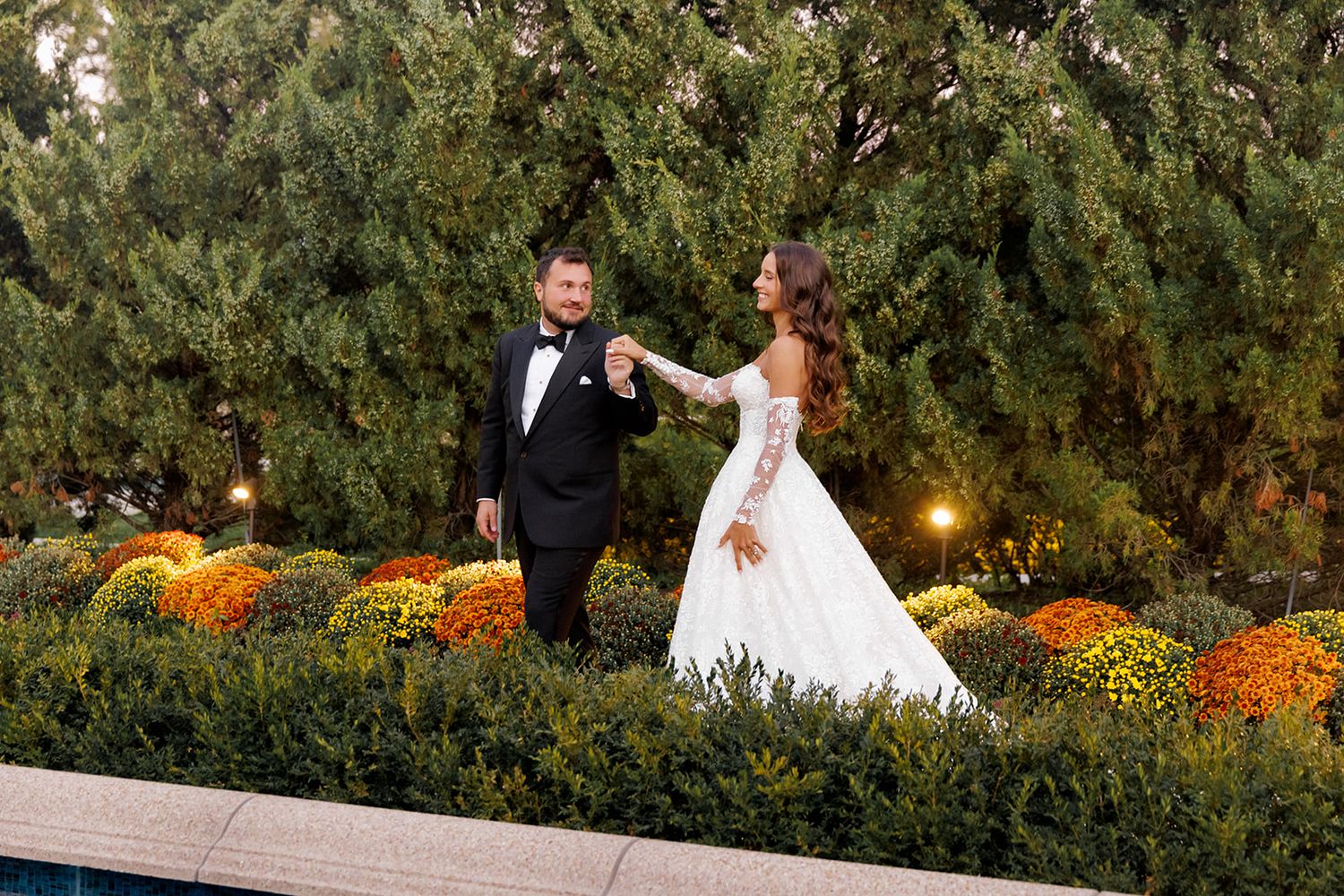 Bride and groom walk hand-in-hand among autumn chrysanthemums and evergreen trees at dusk.