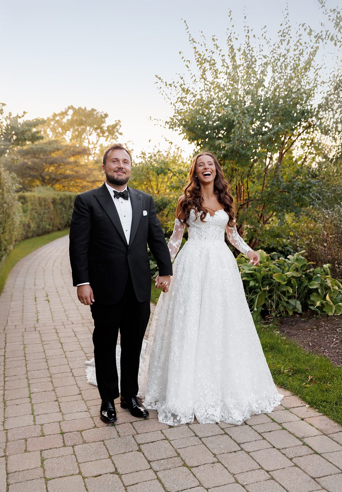 Joyful bride in lace long-sleeve wedding gown walks with tuxedo-clad groom along a garden pathway at sunset.