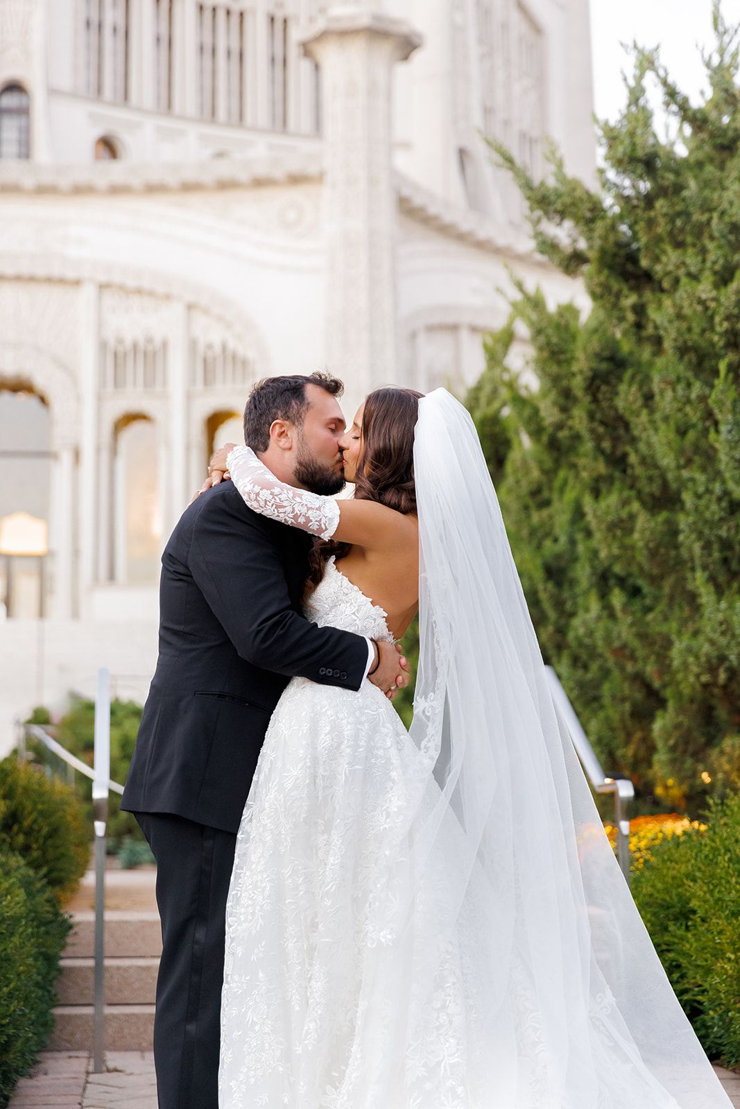 Bride and groom share a romantic kiss outdoors, with bride in a sparkling white gown and veil near an elegant building.