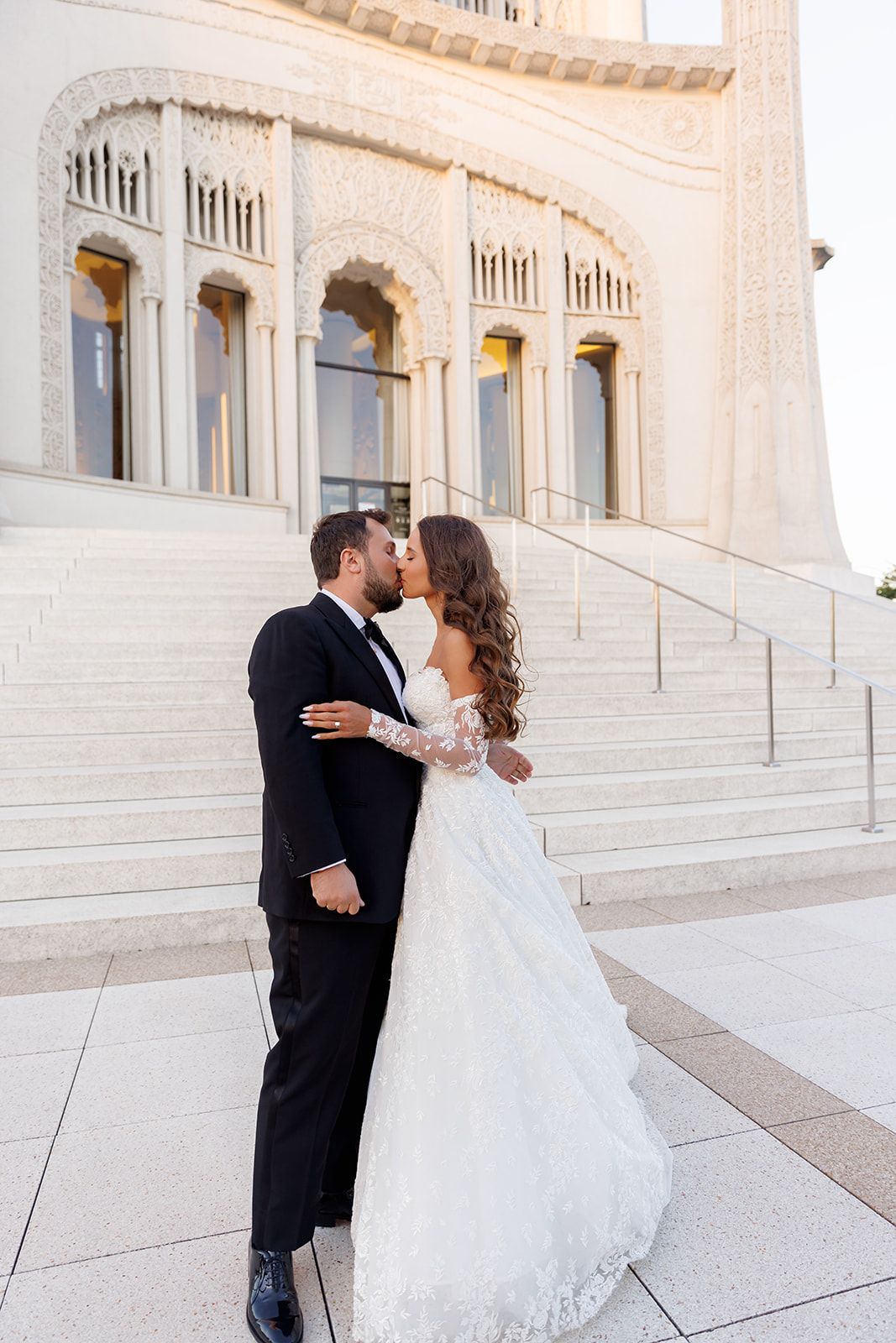 Bride and groom share a kiss on the steps of a stunning white architectural wedding venue.