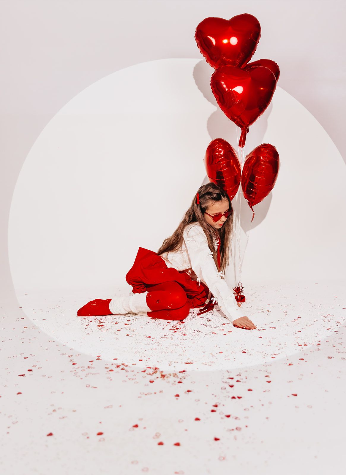 Girl in red outfit sitting on floor with red heart balloons and confetti on white background.