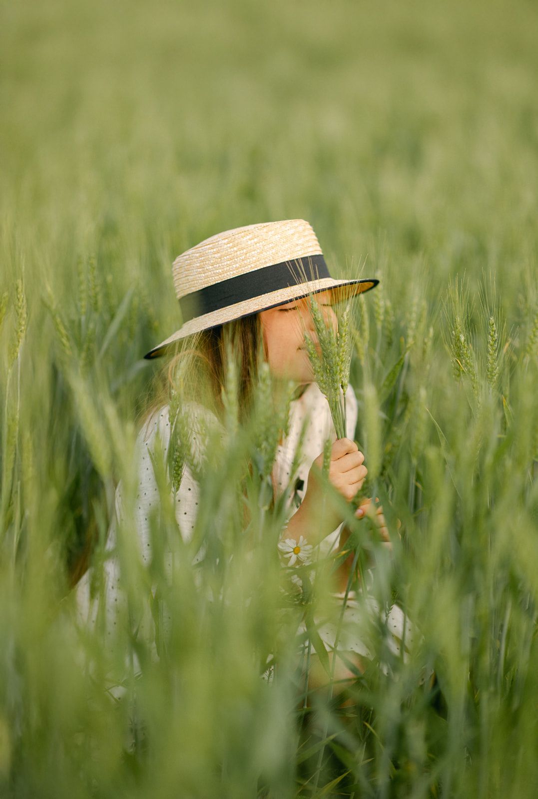 A young girl wearing a straw hat crouches among tall green wheat stalks, holding fresh grain in a field.
