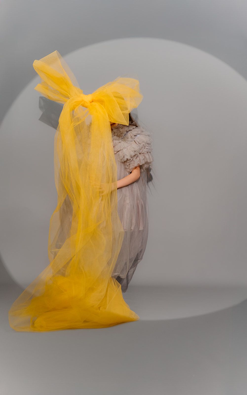A model wearing a ruffled gray gown with a dramatic oversized yellow tulle bow and veil on a gray background.