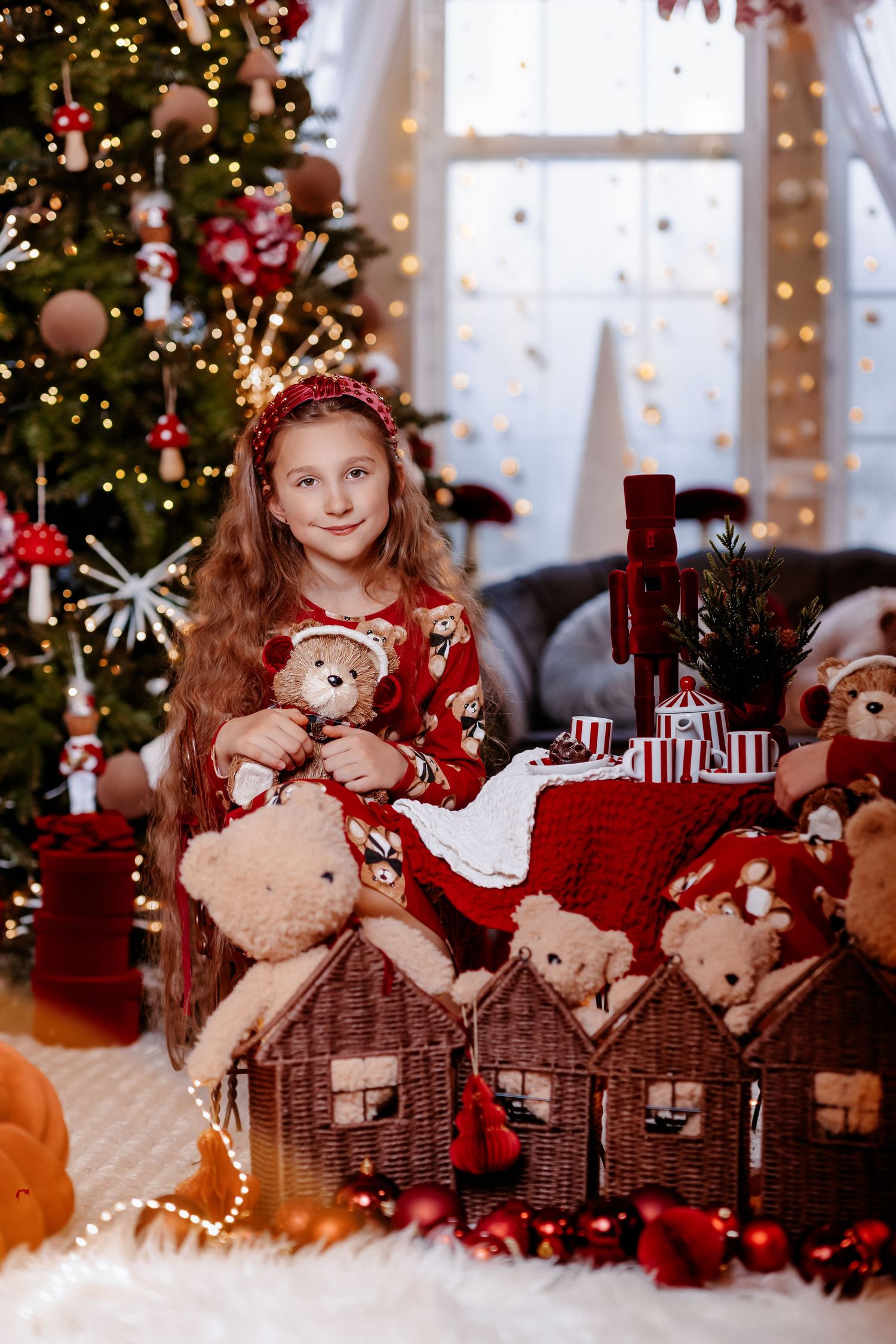 A smiling girl in red pajamas sits among wicker houses and teddy bears in front of a Christmas tree.