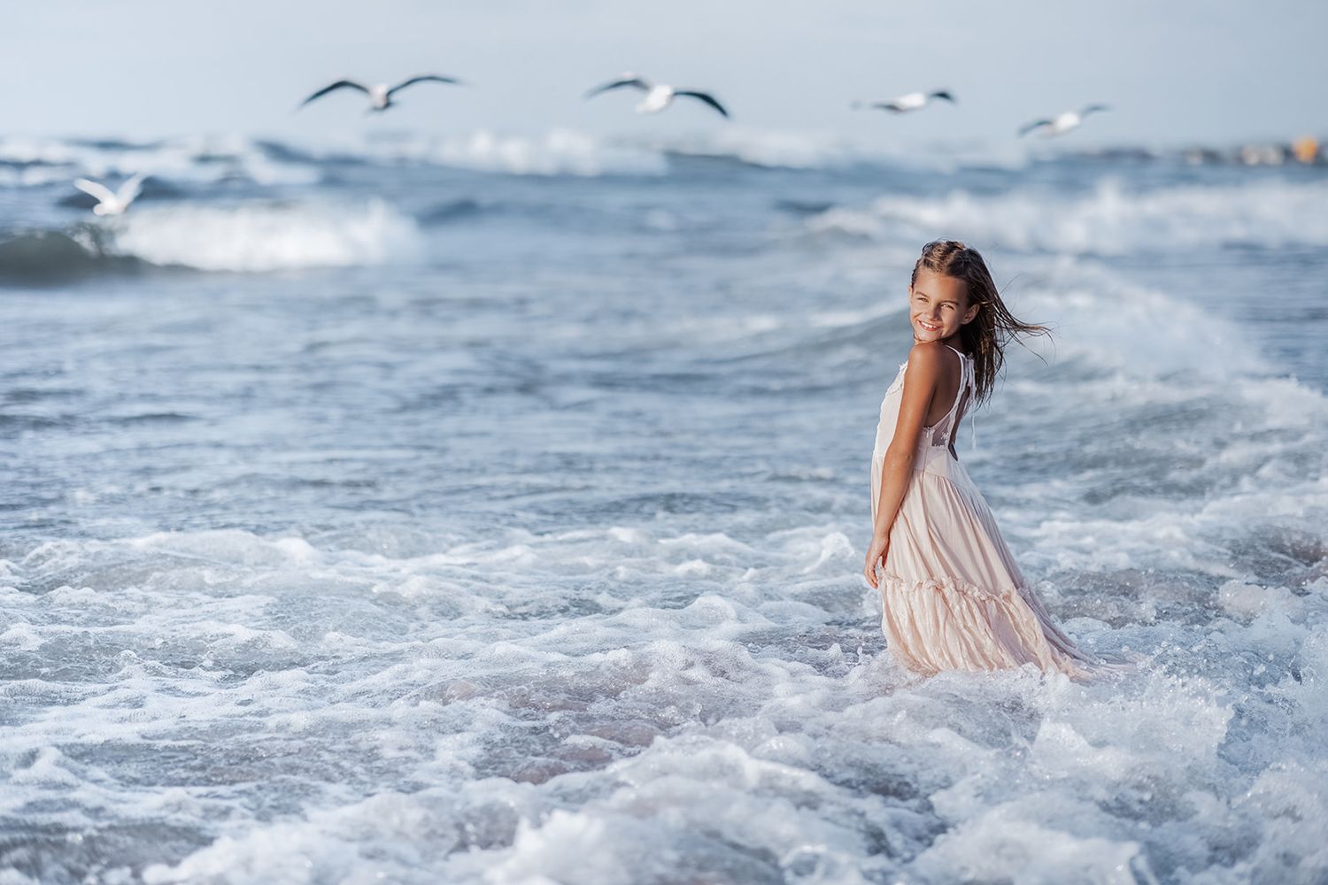 Young woman in pink dress standing in ocean waves with seagulls flying overhead on a cloudy beach day.