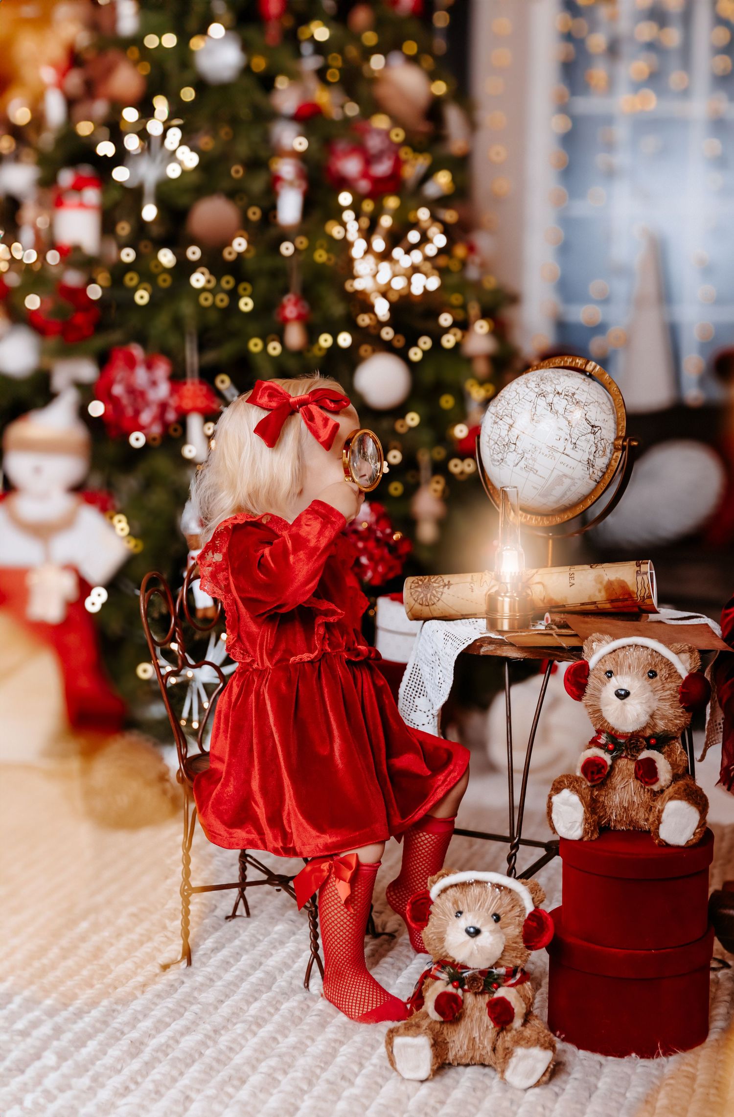 A little girl in a red velvet dress sits before a decorated Christmas tree with teddy bears and a globe nearby.