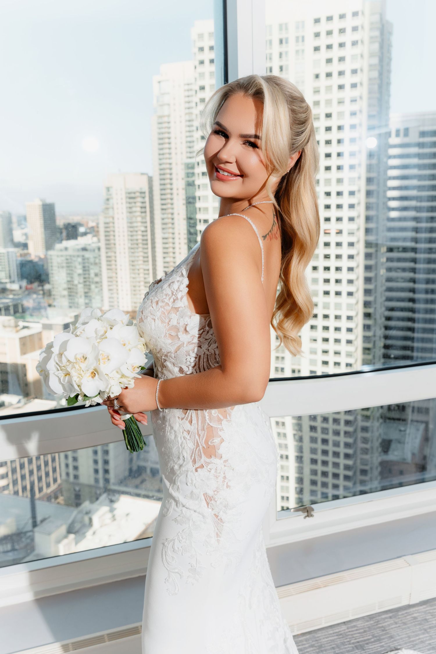 Smiling blonde bride in lace wedding dress holding white orchid bouquet before urban skyline windows.