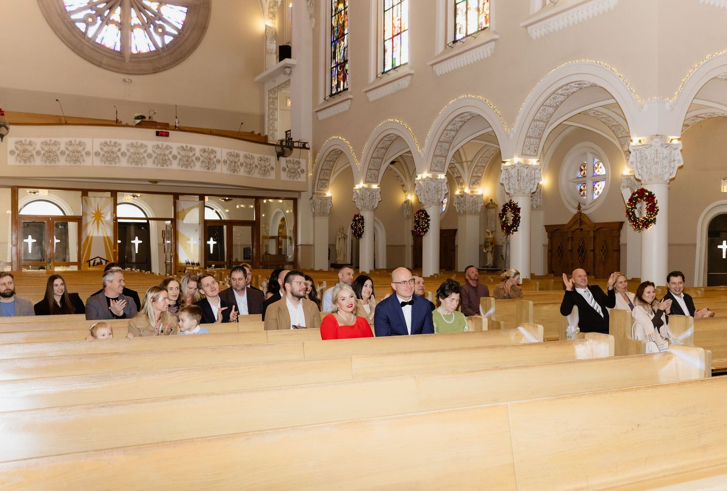 Large group of wedding guests seated in light wood church pews beneath a stunning rose window and arched columns.