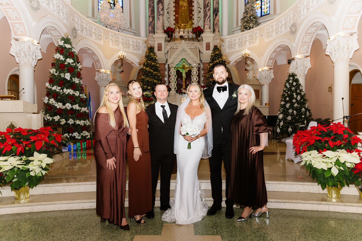 Bridal party of six stands at church altar adorned with poinsettias and Christmas trees during winter wedding ceremony.