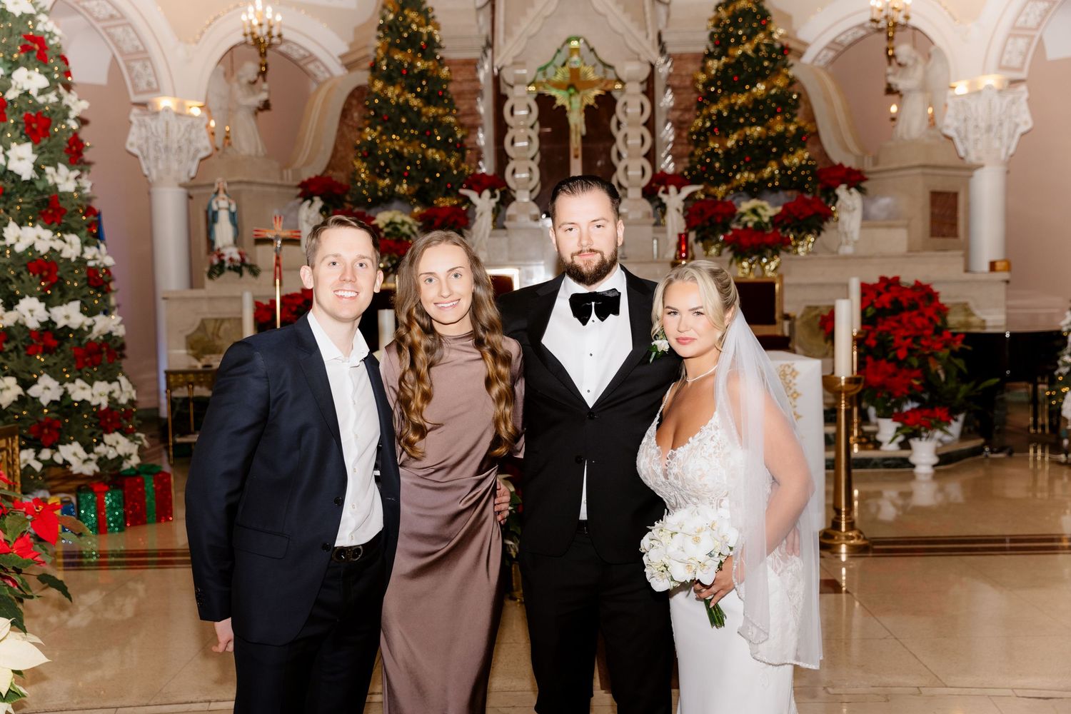 Bride and groom pose with two guests inside a Christmas-decorated church with poinsettias and trees.
