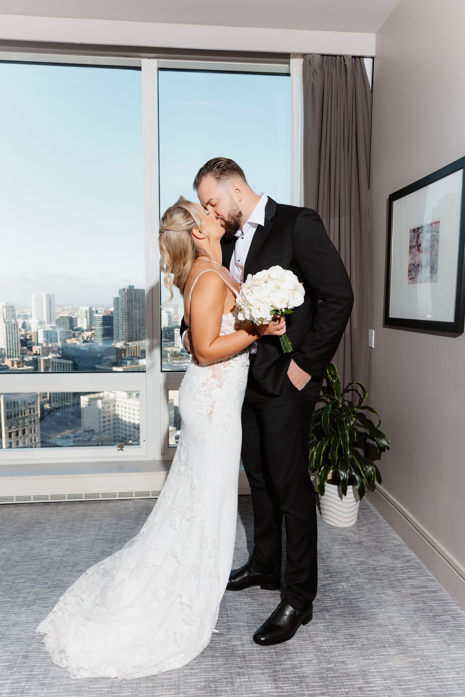 Bride in lace gown and groom in black suit share a romantic kiss holding white bouquet with city skyline behind them.