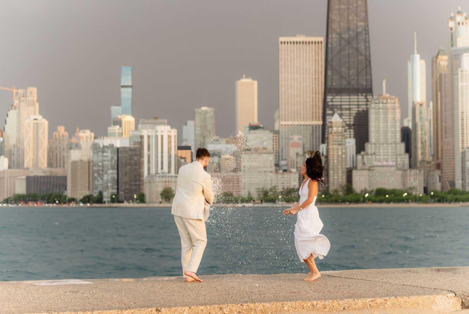 Couple in white outfits playfully splashing water by Chicago lakefront skyline at sunset.