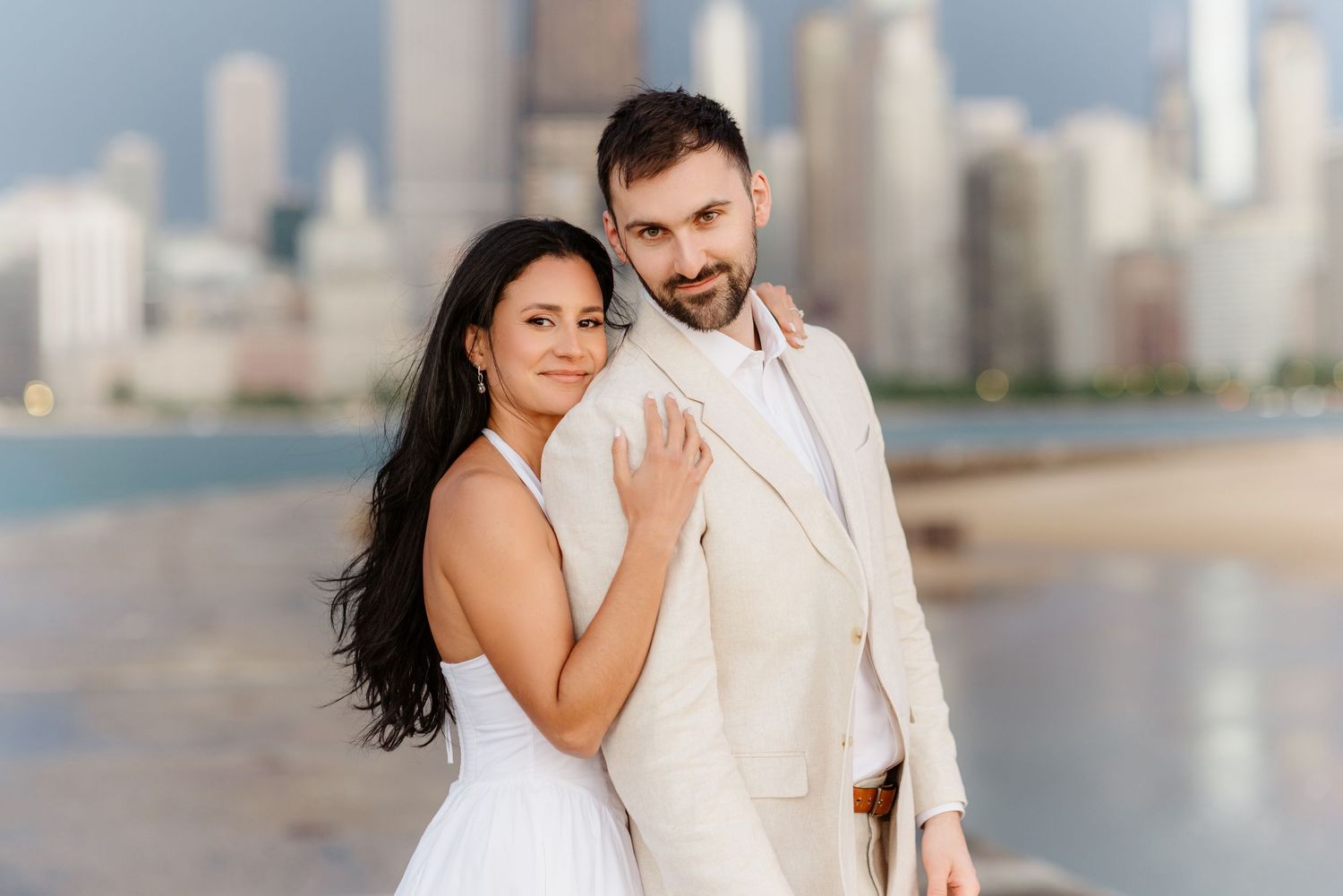 Elegant couple posing together on Chicago beach, bride in white halter dress and groom in cream suit.