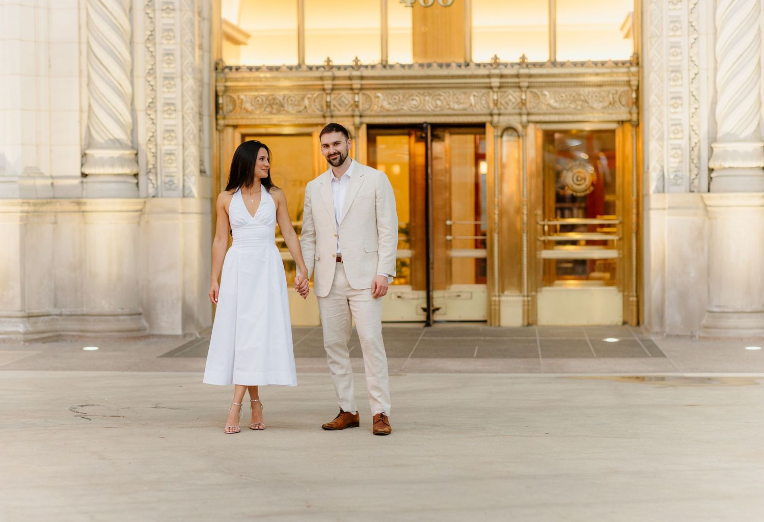 Couple in white dress and beige suit holding hands outside ornate building entrance.
