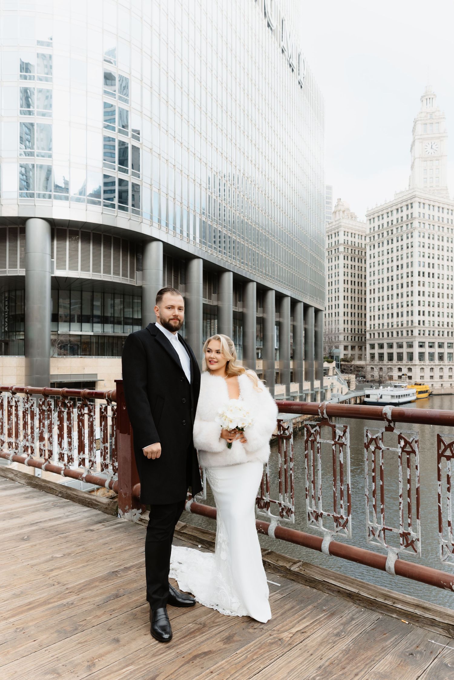 Bride and groom pose on Chicago Riverwalk boardwalk with city skyline behind them.