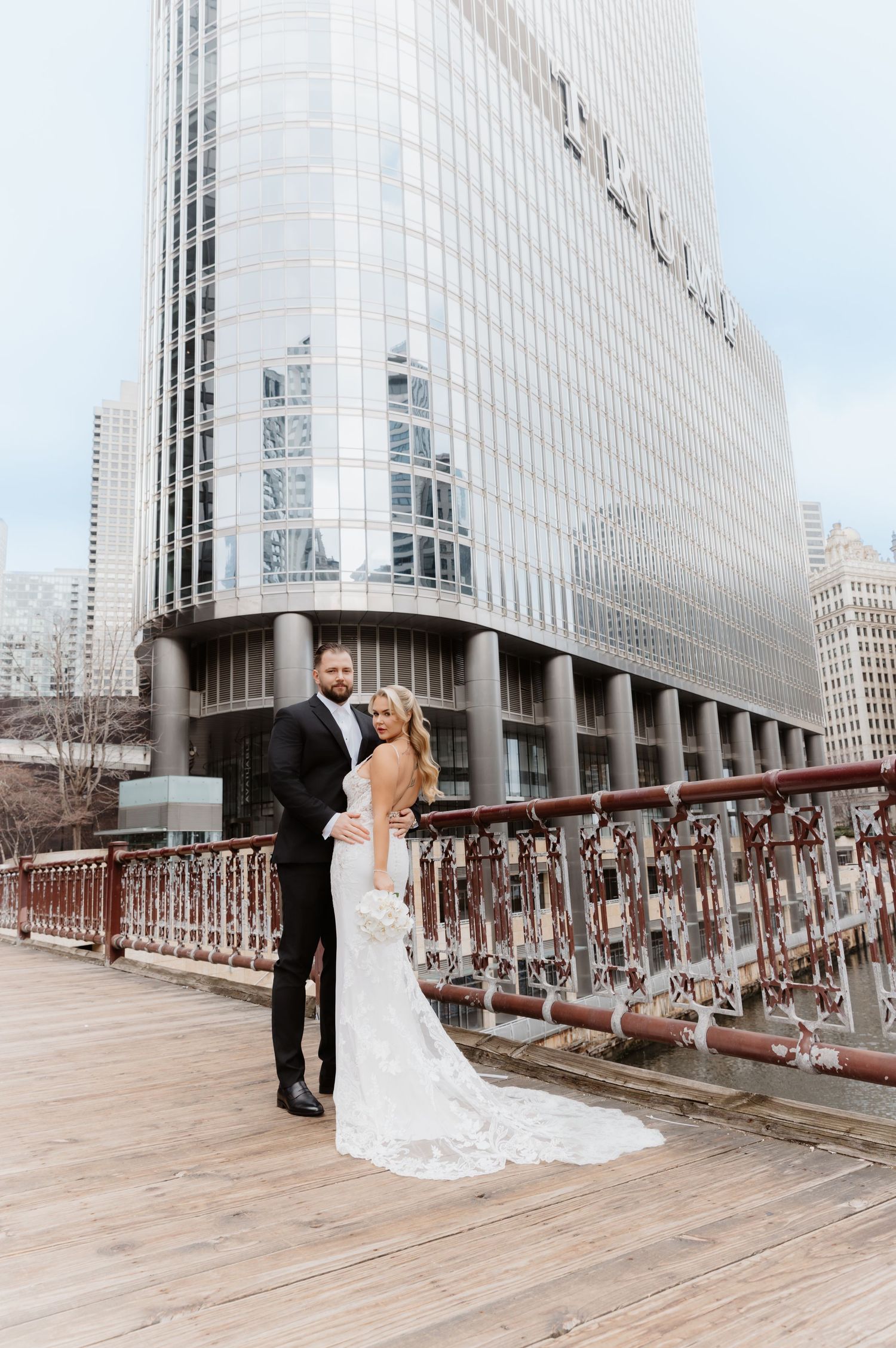 Wedding couple embraces on Chicago riverwalk boardwalk with towering glass skyscraper in background.
