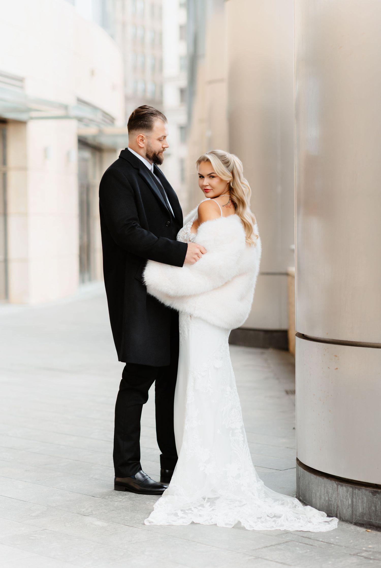 Bride in white lace gown with fur wrap and groom in black tuxedo share romantic urban moment.