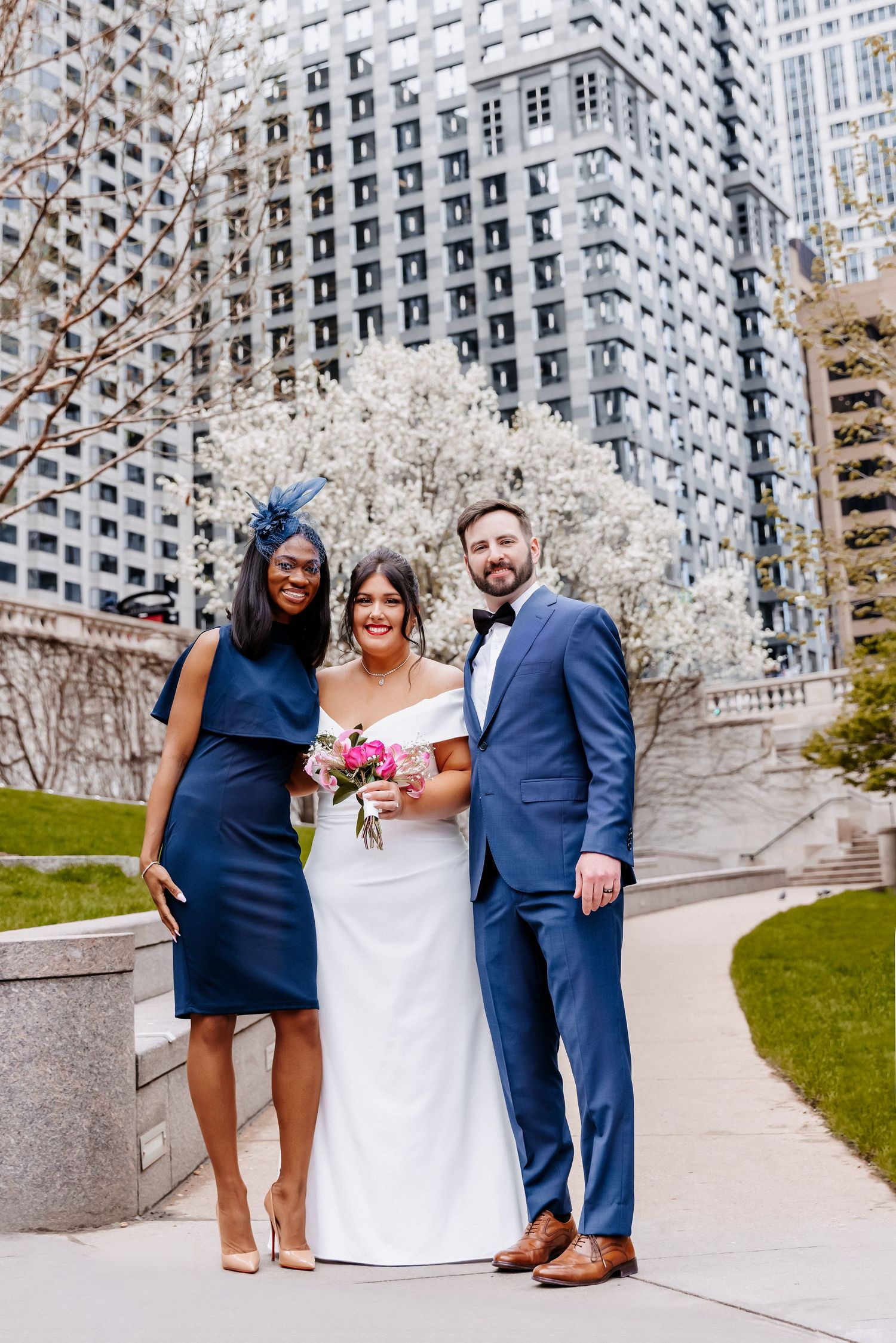 Bride in white off-shoulder gown poses with groom in blue suit and bridesmaid in navy dress.