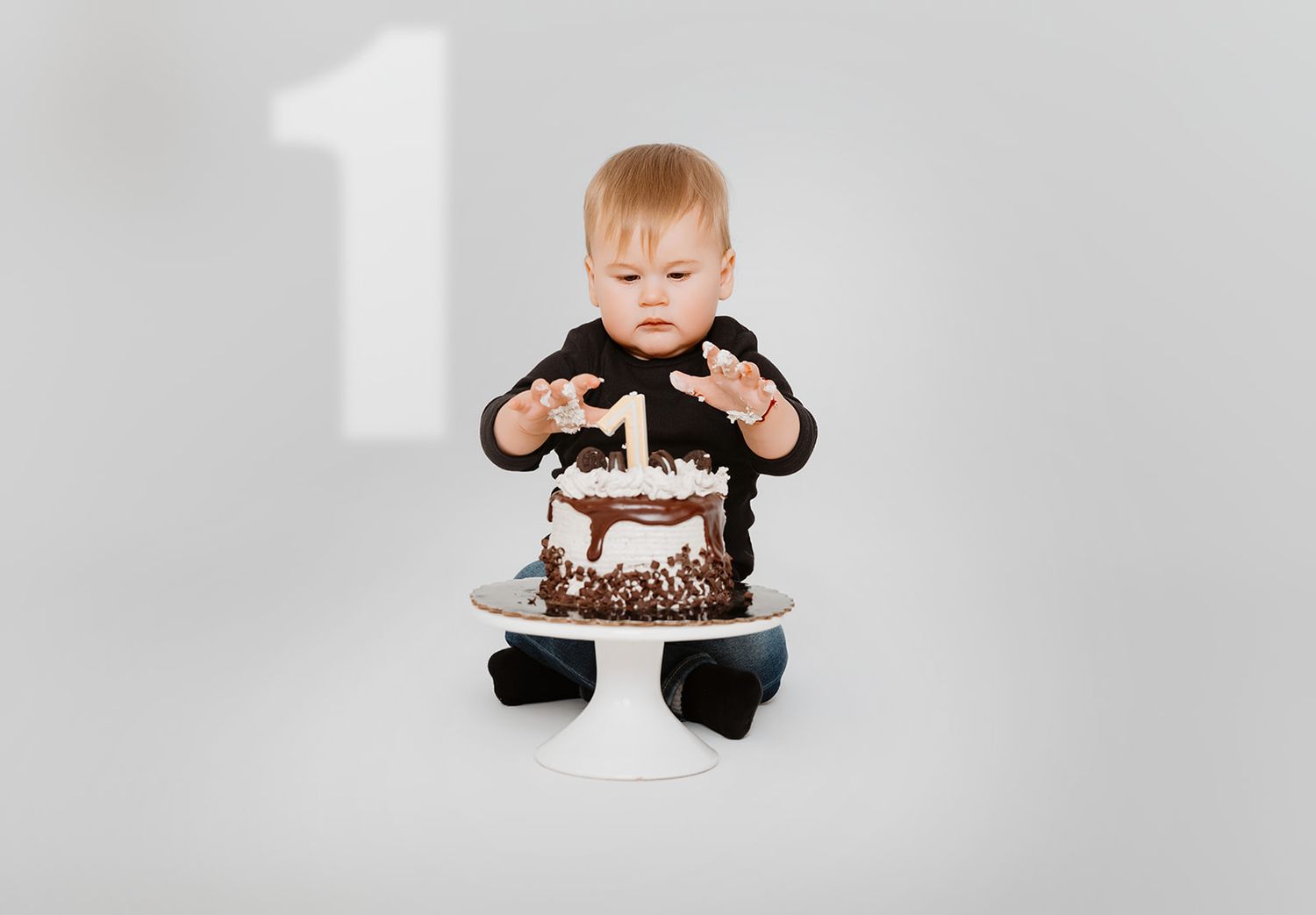 Toddler in black outfit reaching for chocolate birthday cake with number 1 on white background.