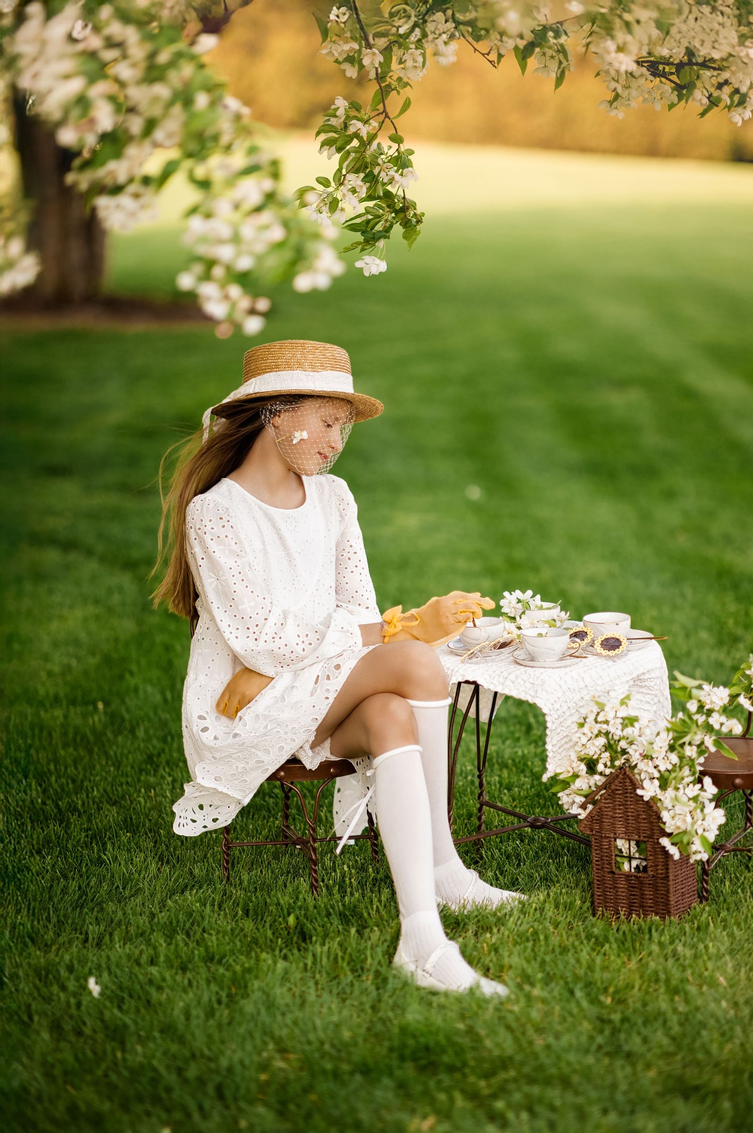 Young girl in white lace dress and straw hat enjoys a tea party outdoors under a blooming apple tree.