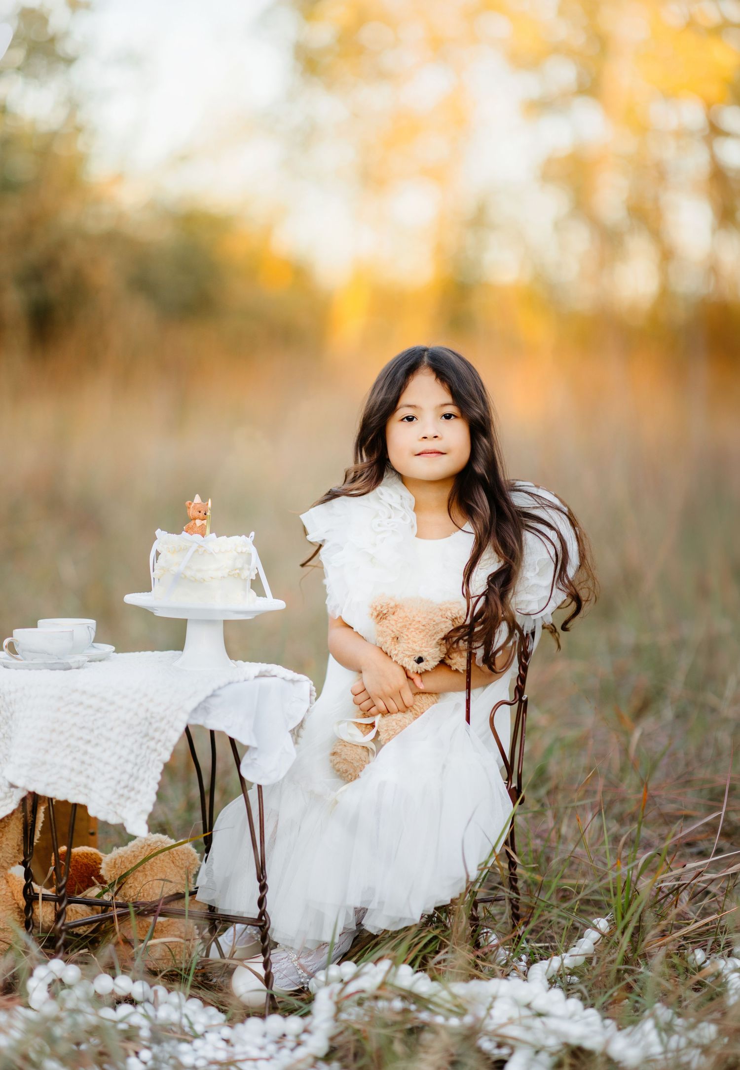 A young girl in a white dress holds a teddy bear outdoors, seated near a birthday cake in a golden field.