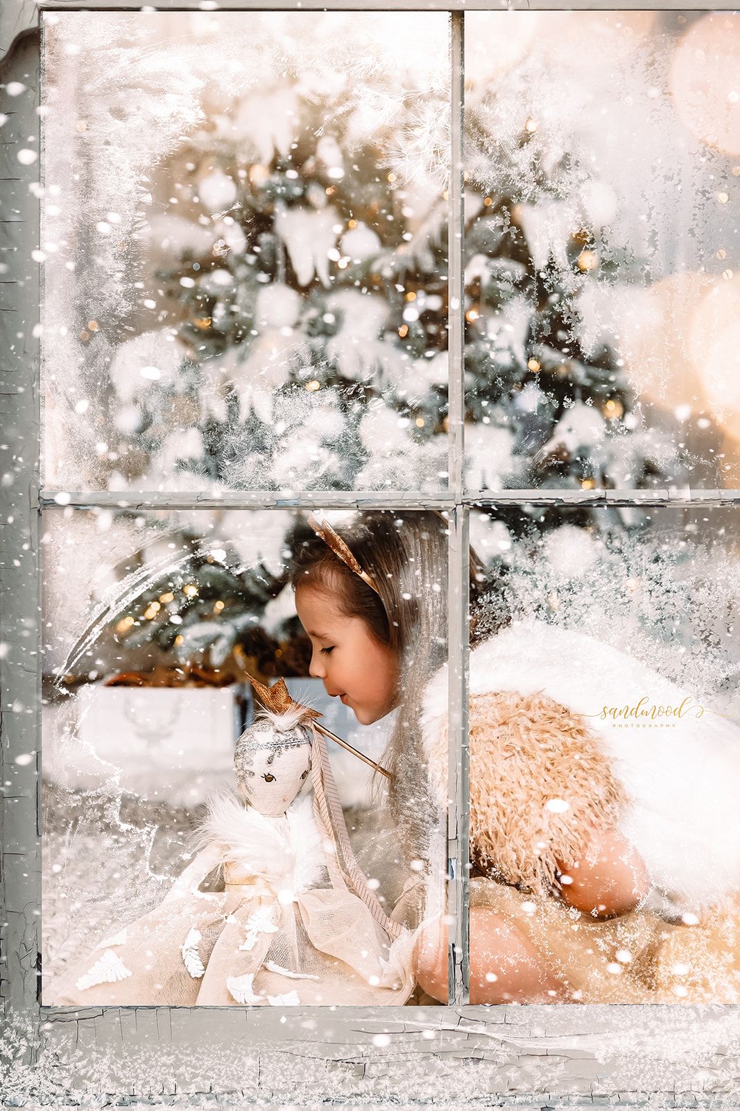 Young girl looking at doll and teddy bear through snow-frosted window with Christmas tree in background.