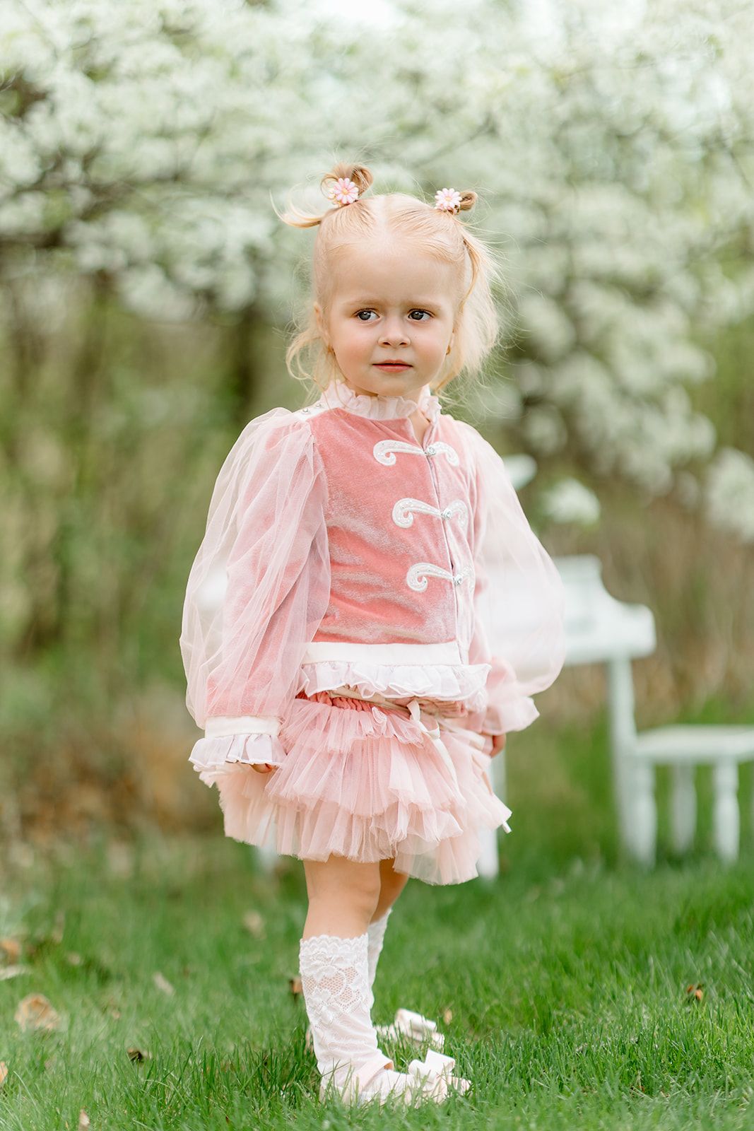 Adorable blonde toddler girl in pink tutu dress and white boots standing on green grass near blooming trees.