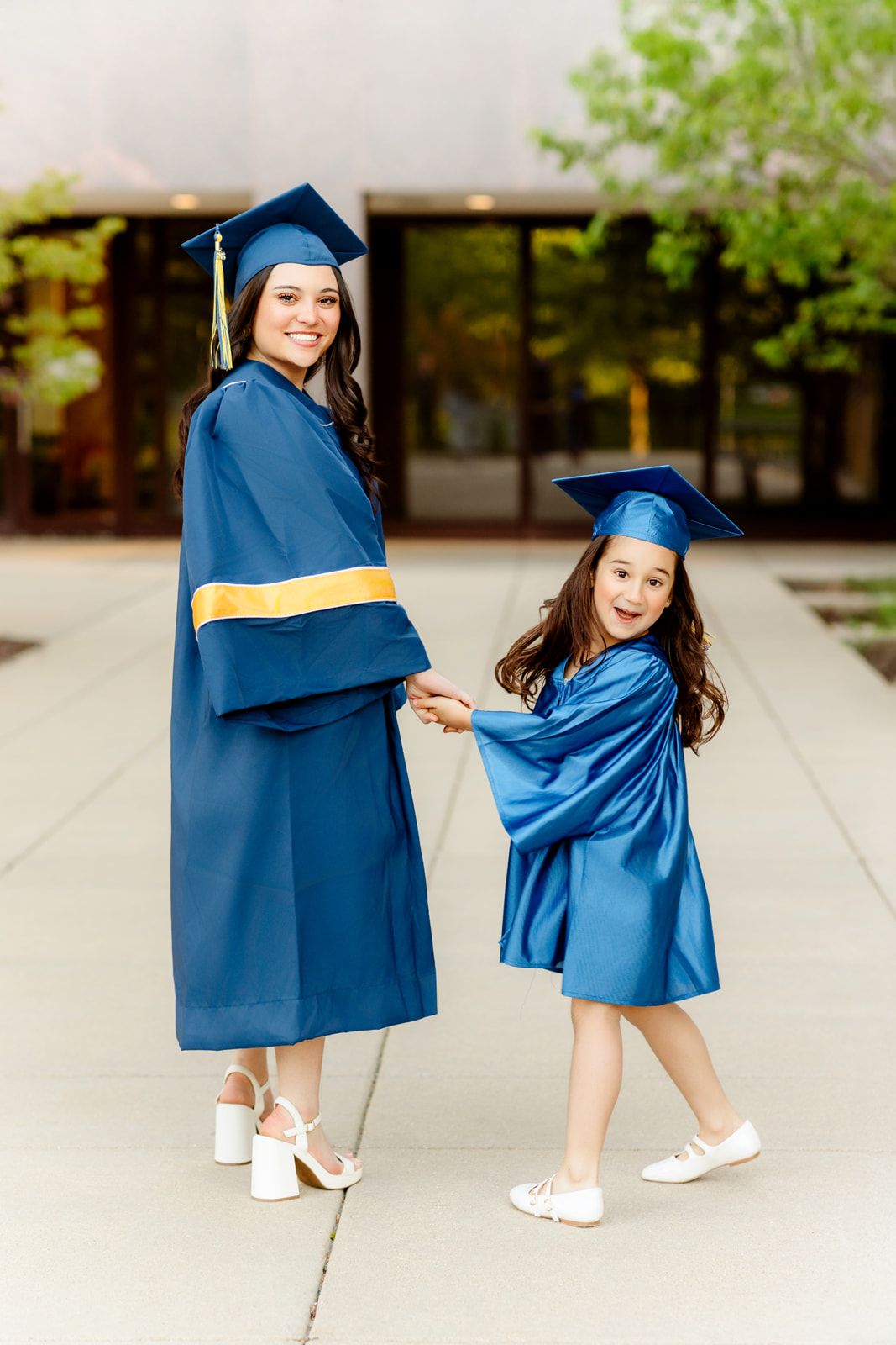 Smiling graduate in navy gown holding hands with young girl in matching blue graduation attire outdoors.