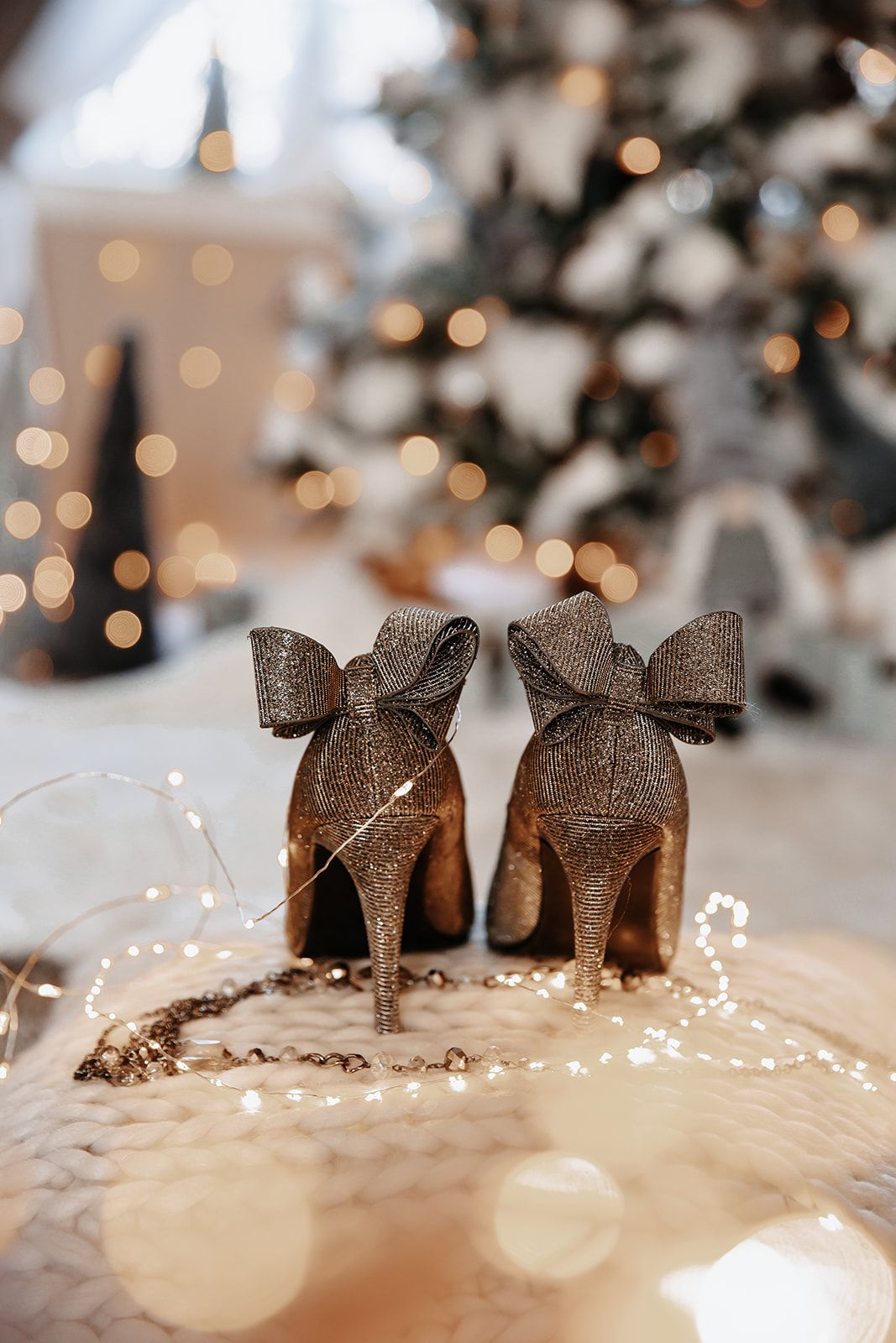 Glittery gold high heels with bow detail displayed on white knit surface with bokeh Christmas tree lights in background.