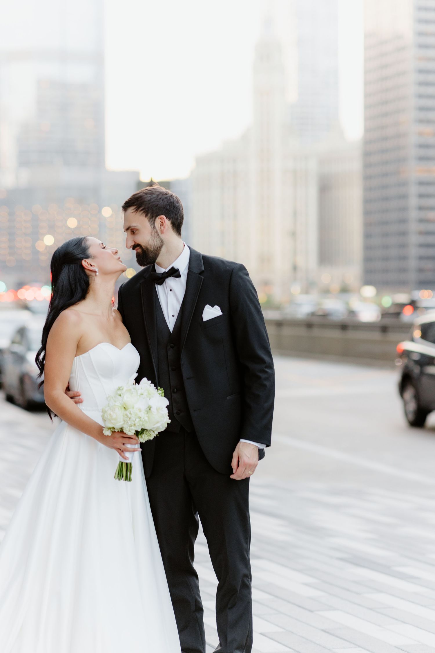 Bride and groom share a romantic almost-kiss on a city street, groom in black tuxedo holding white bouquet.
