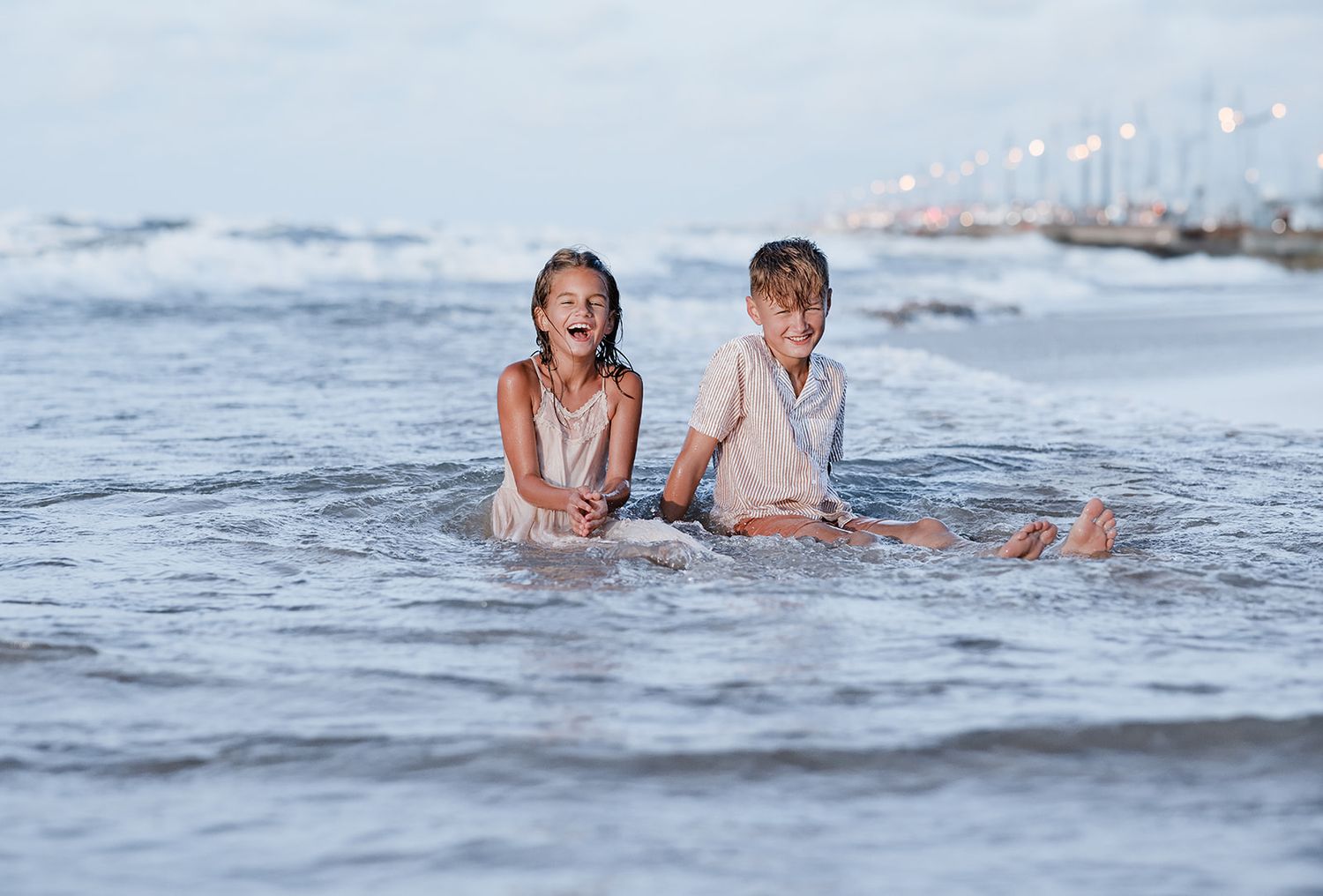 Two happy children sitting in ocean waves on a sandy beach, laughing and playing in the shallow water at dusk.