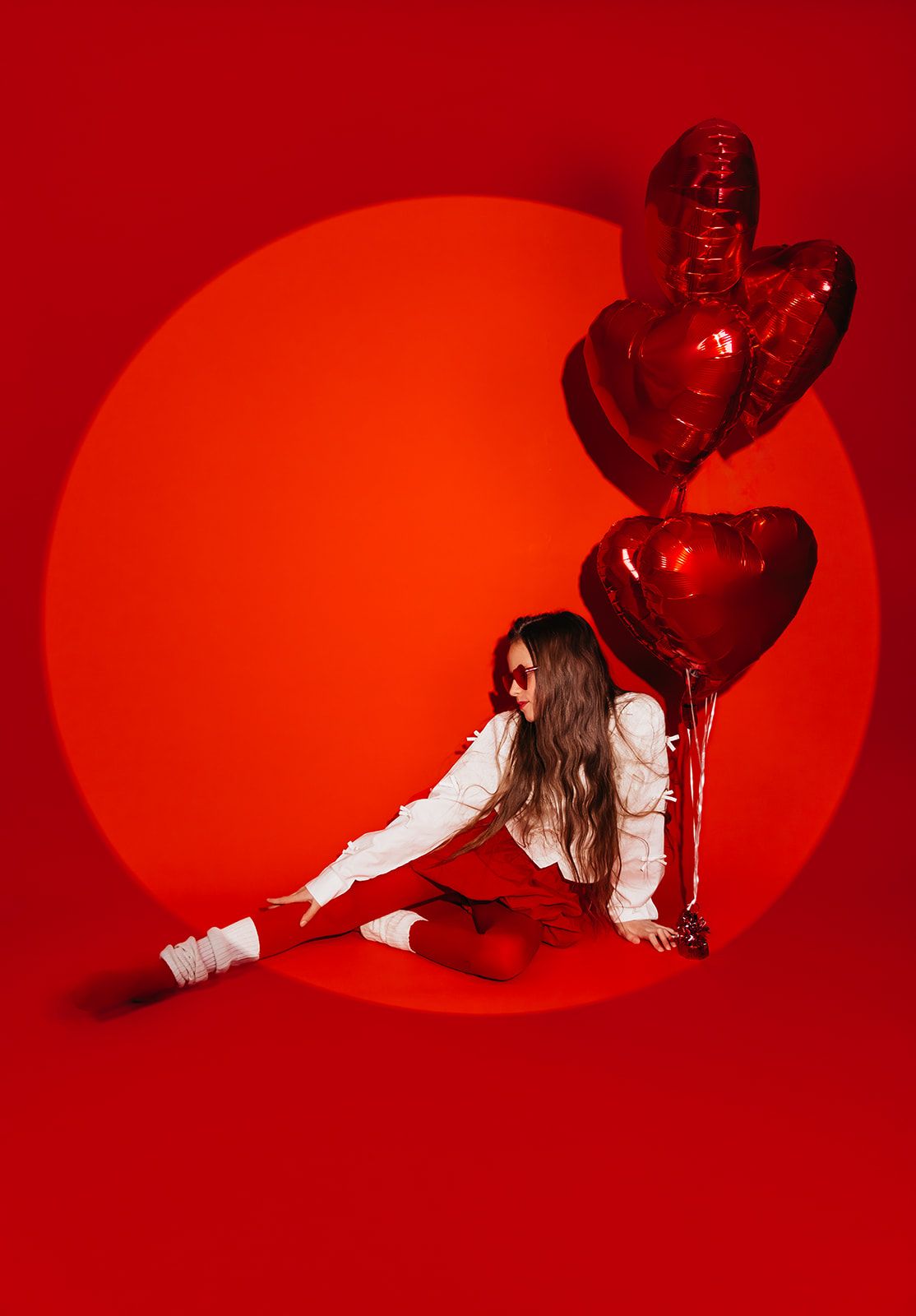 Woman in red outfit sitting on floor with red heart-shaped foil balloons against bold red background.