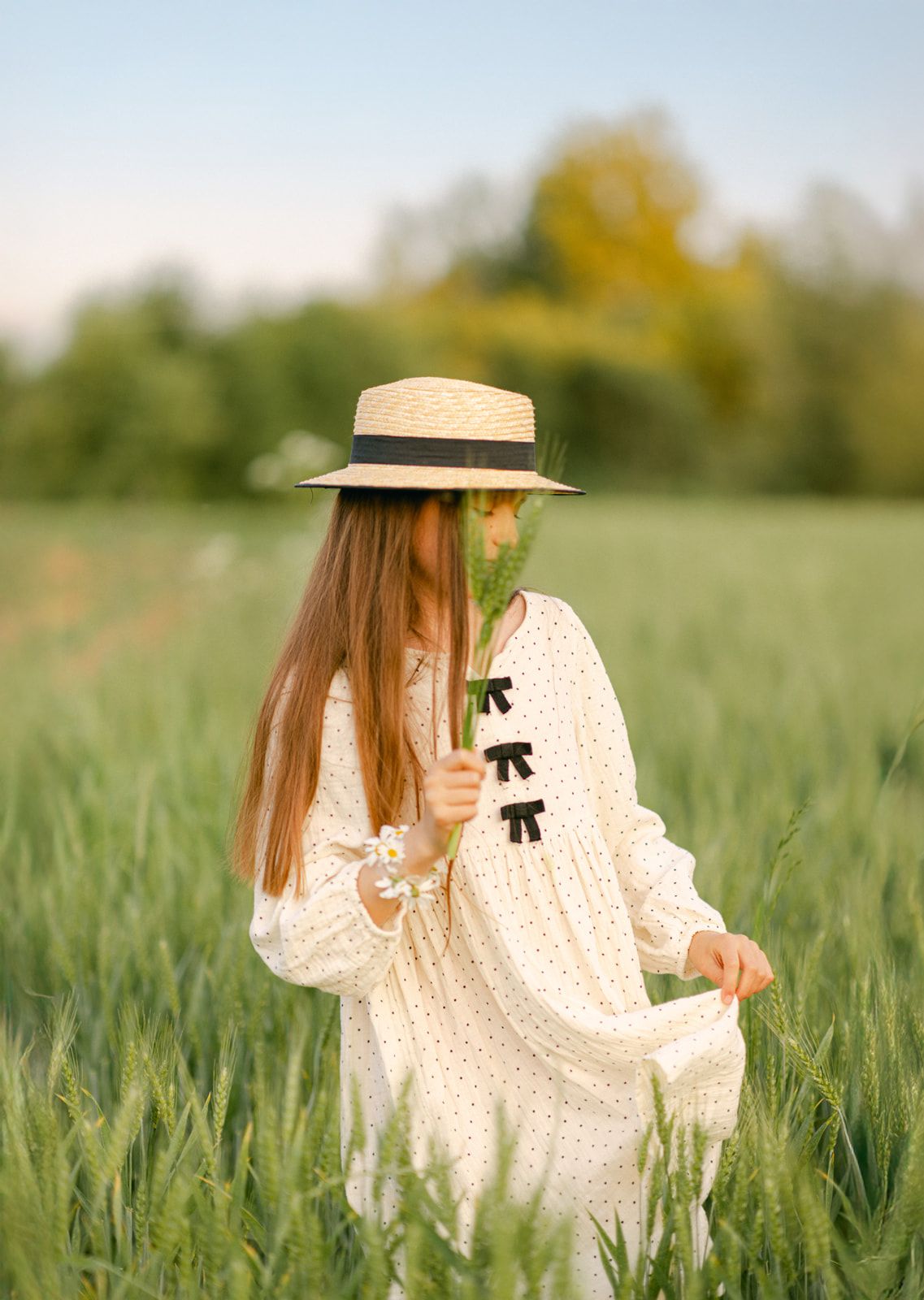 A young girl in a white polka dot dress and straw hat holds green wheat stalks in a lush field.