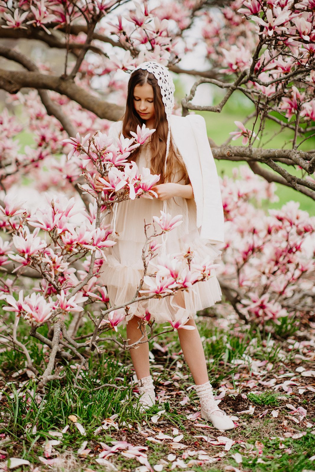Young girl in cream dress and white bonnet holding pink magnolia flowers while standing among blooming spring trees.