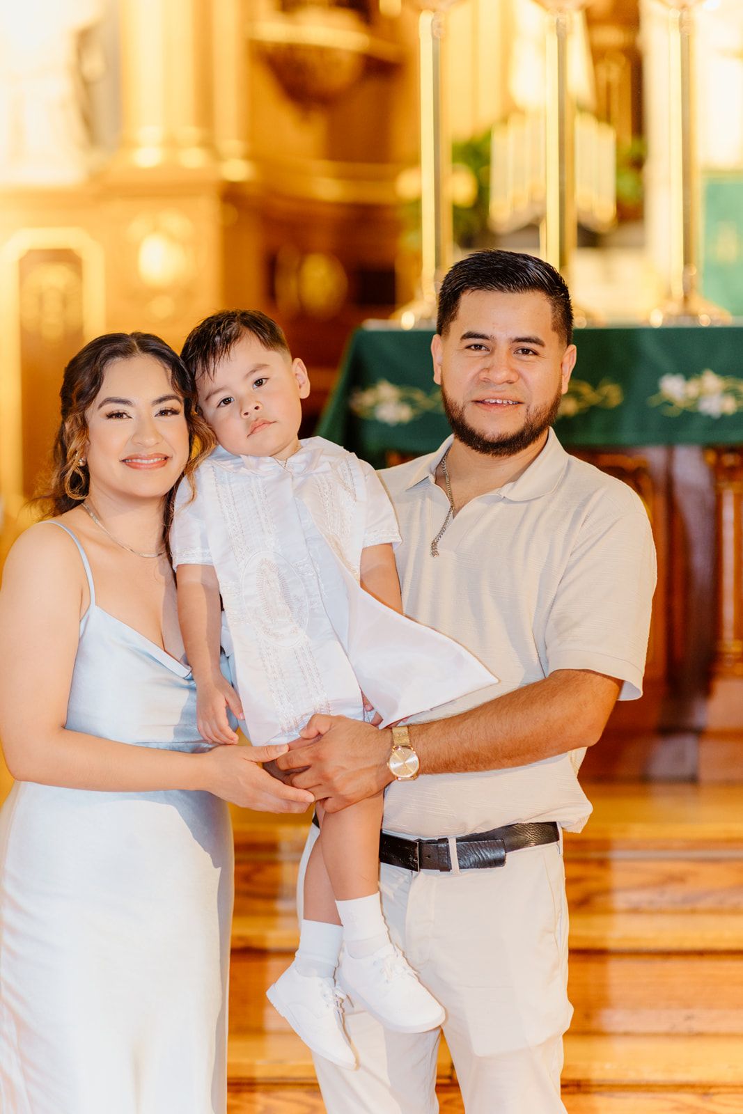 Smiling parents holding toddler in white baptism outfit inside ornate church interior.