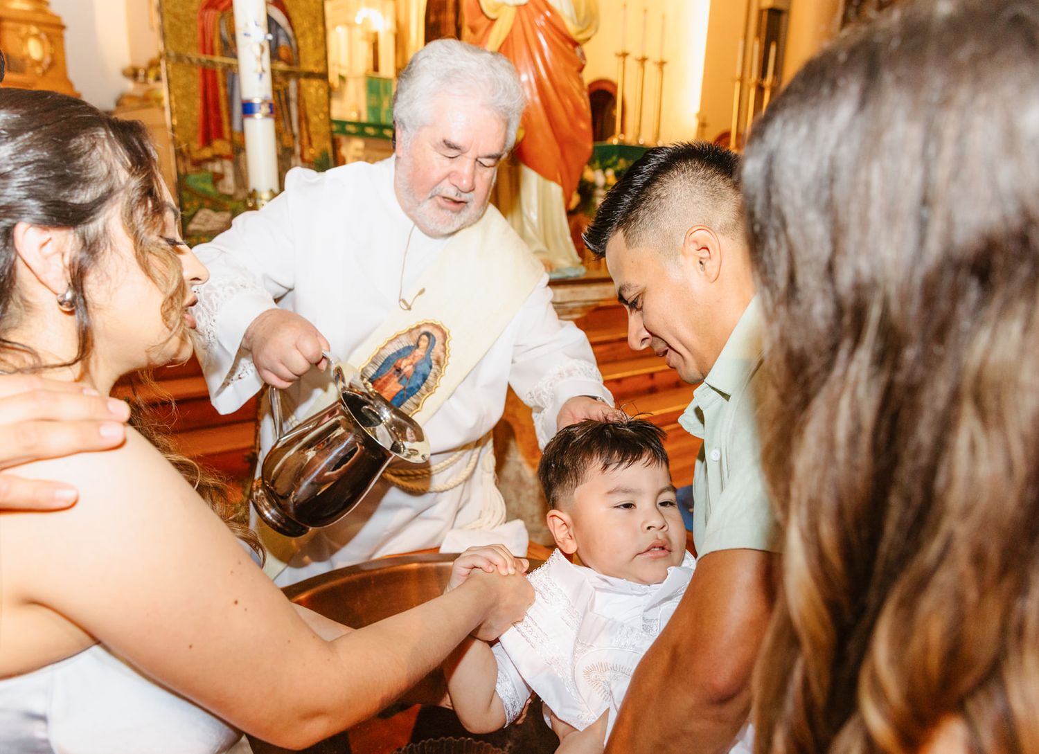 Priest performing Catholic baptism ceremony, pouring holy water over infant held by parents in ornate church.
