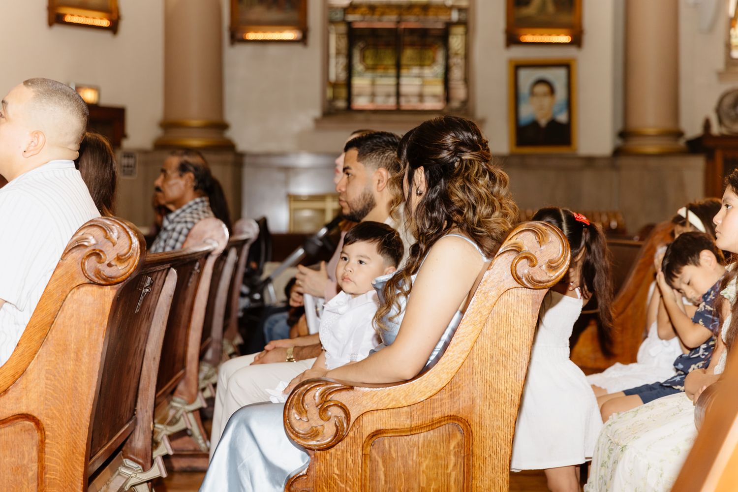 Parents holding a baby in church pews during a religious ceremony, surrounded by family members in formal attire.