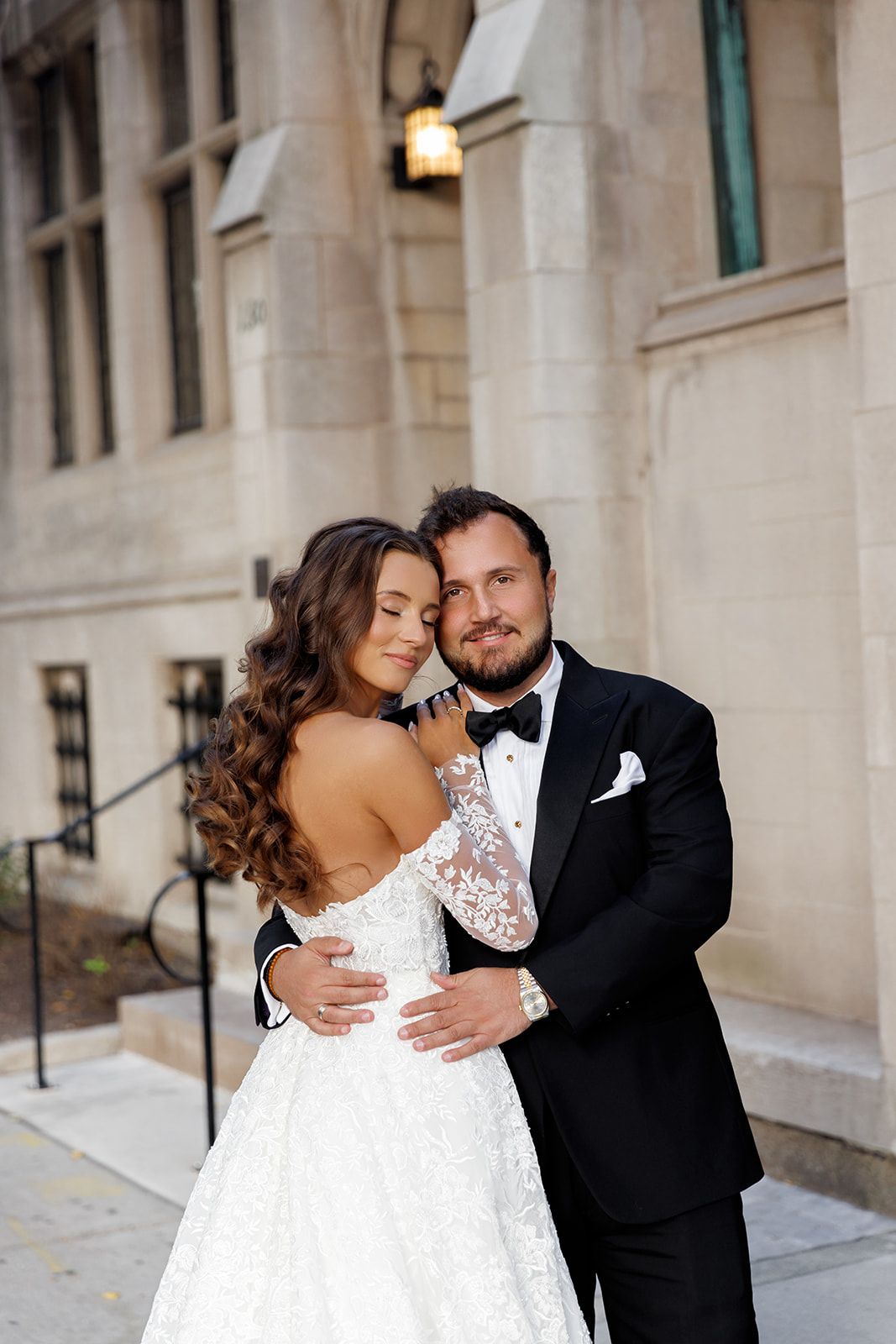 Bride in lace gown and groom in black tuxedo embrace tenderly outside elegant stone building on wedding day.