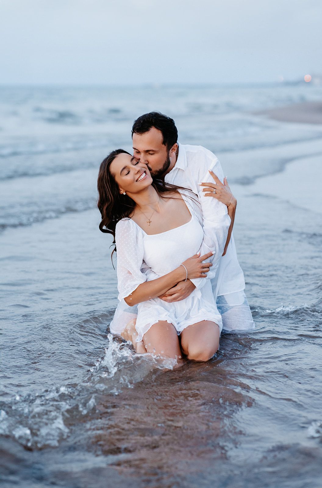 Romantic couple in white outfits embracing in ocean waves on a beach at dusk.
