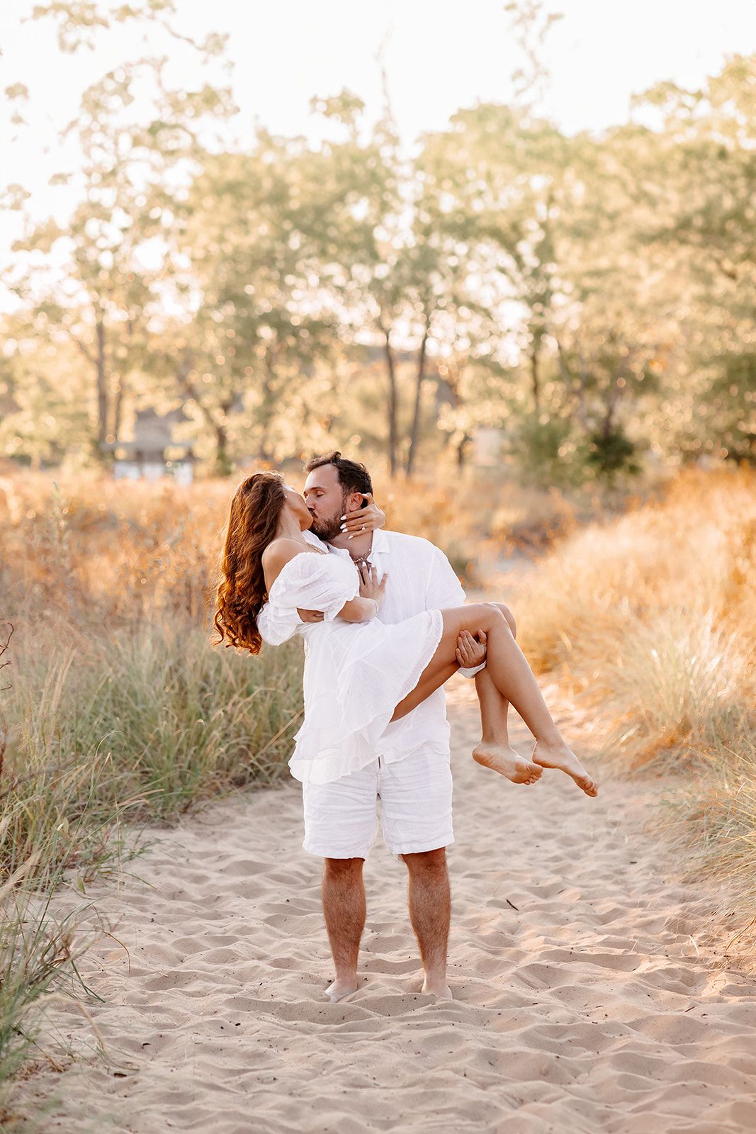 A man in white carries a woman in his arms while kissing on a sandy beach path surrounded by golden grass.