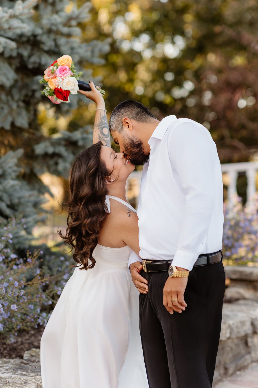 Bride in white dress holds colorful bouquet aloft while kissing tattooed groom in white shirt outdoors.