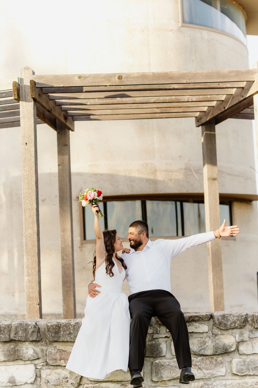 Bride and groom pose romantically by a stone wall under a wooden pergola, she holds a colorful bouquet.