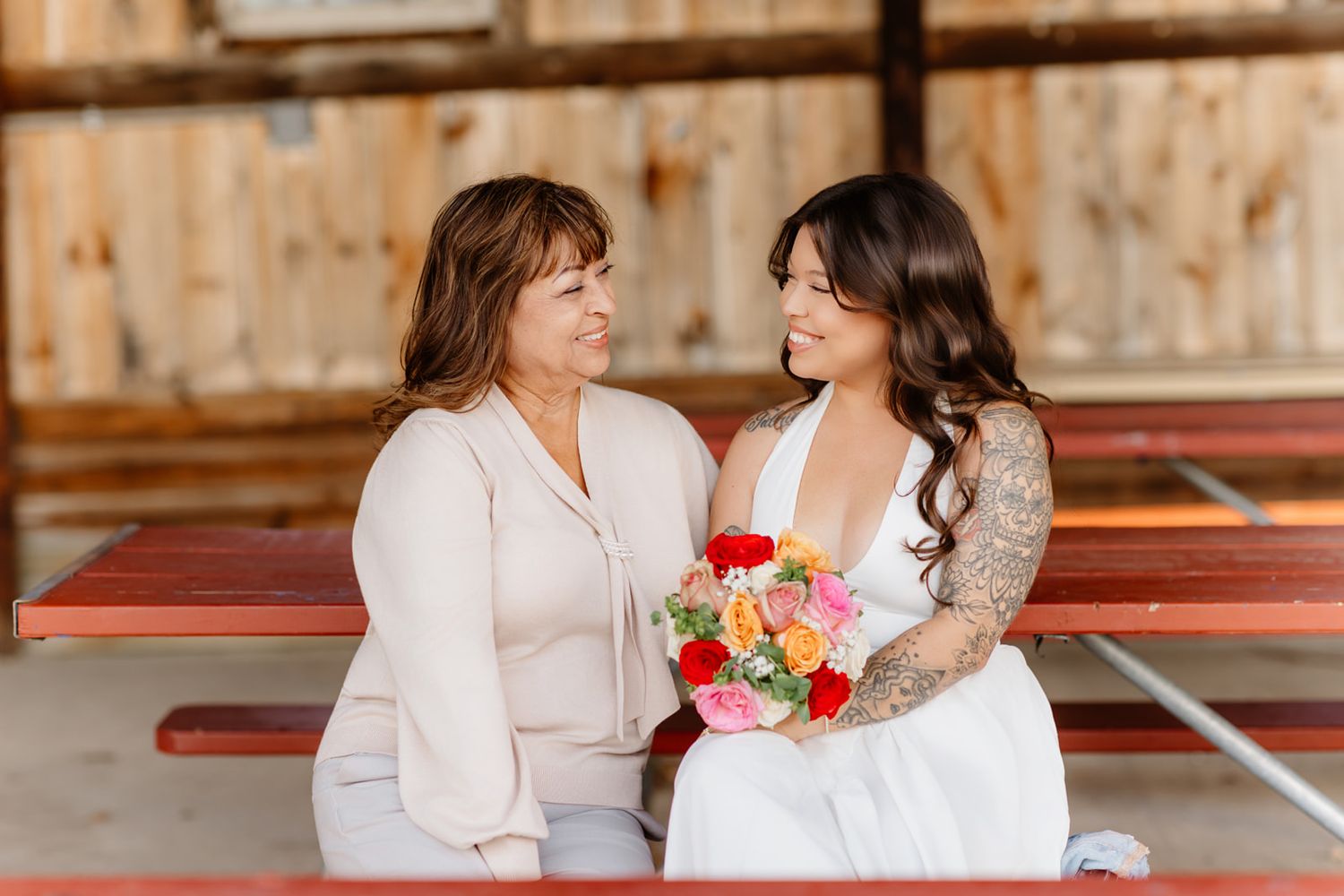 Bride in white dress holding colorful bouquet shares a sweet smile with her mother on a red bench.