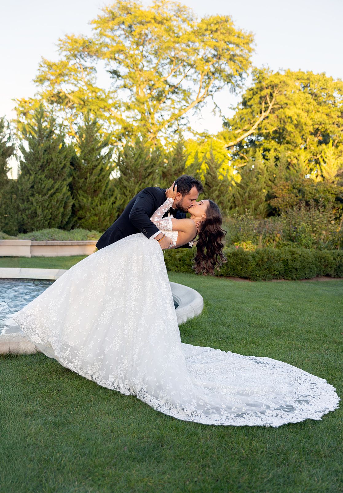 Couple shares a romantic dip kiss on sunlit lawn, bride in lace gown with long train near garden pool.