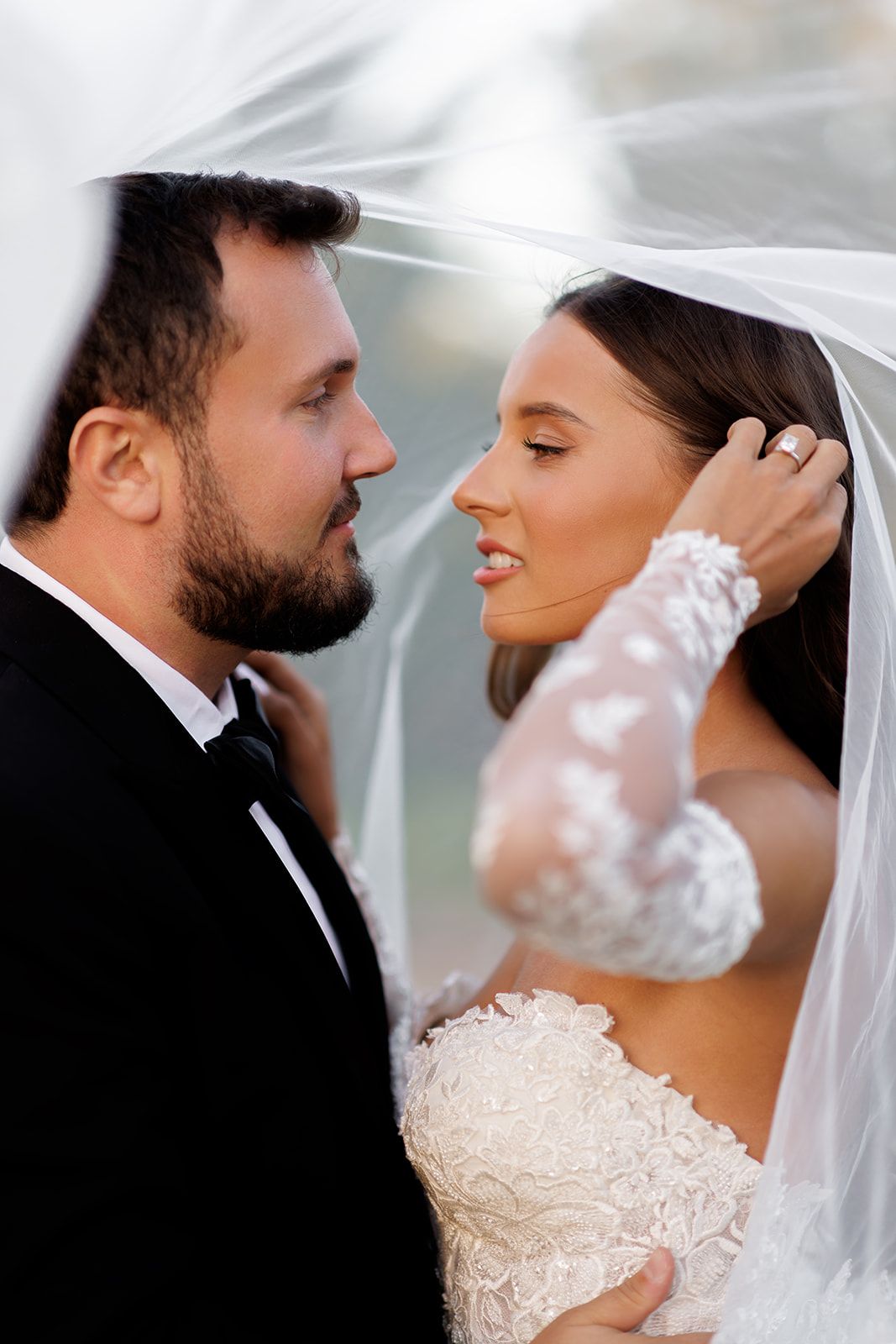 Romantic close-up of bride and groom sharing intimate moment under flowing white wedding veil.