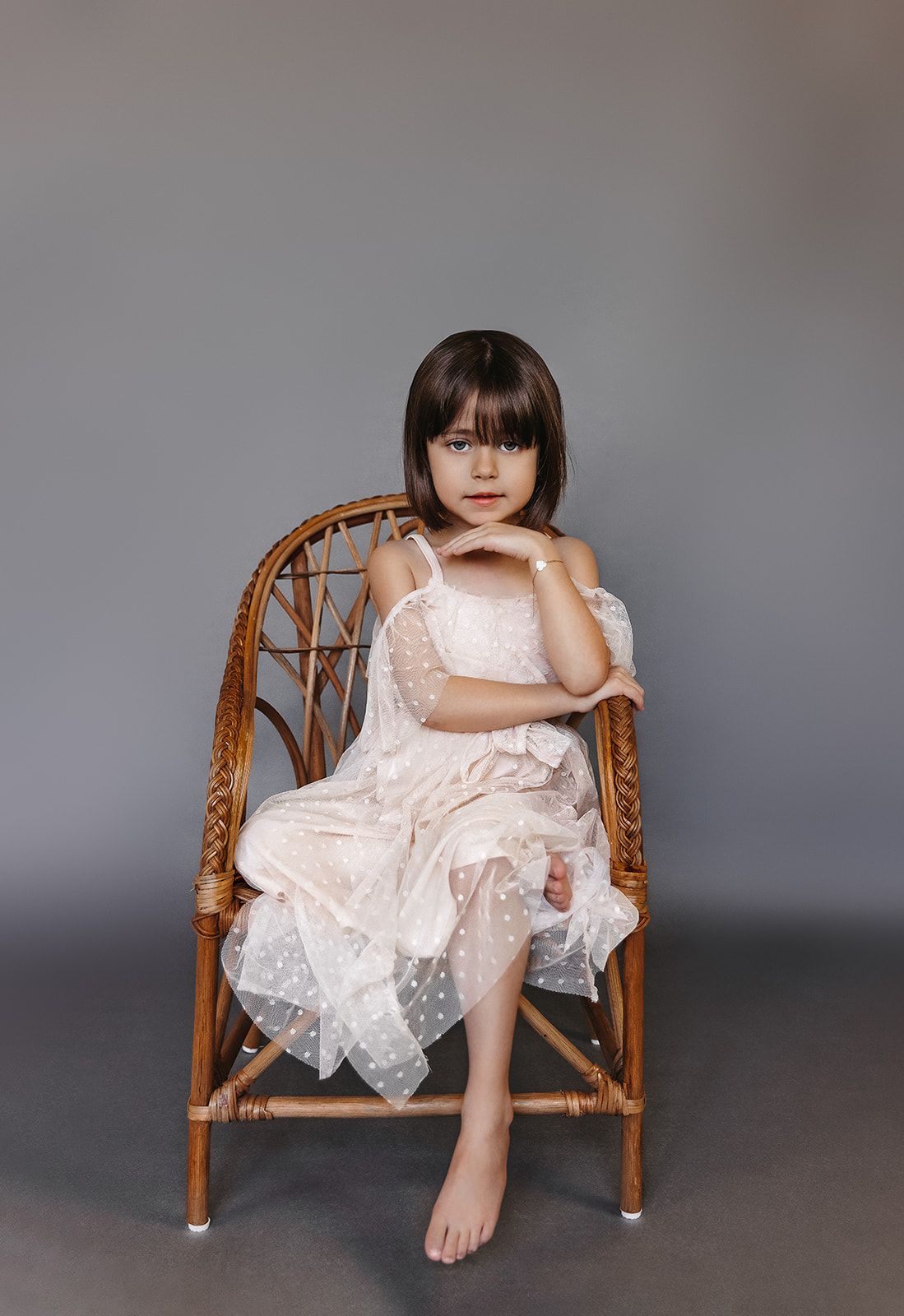 Little girl in white polka dot tulle dress sits barefoot on wicker chair against gray background.