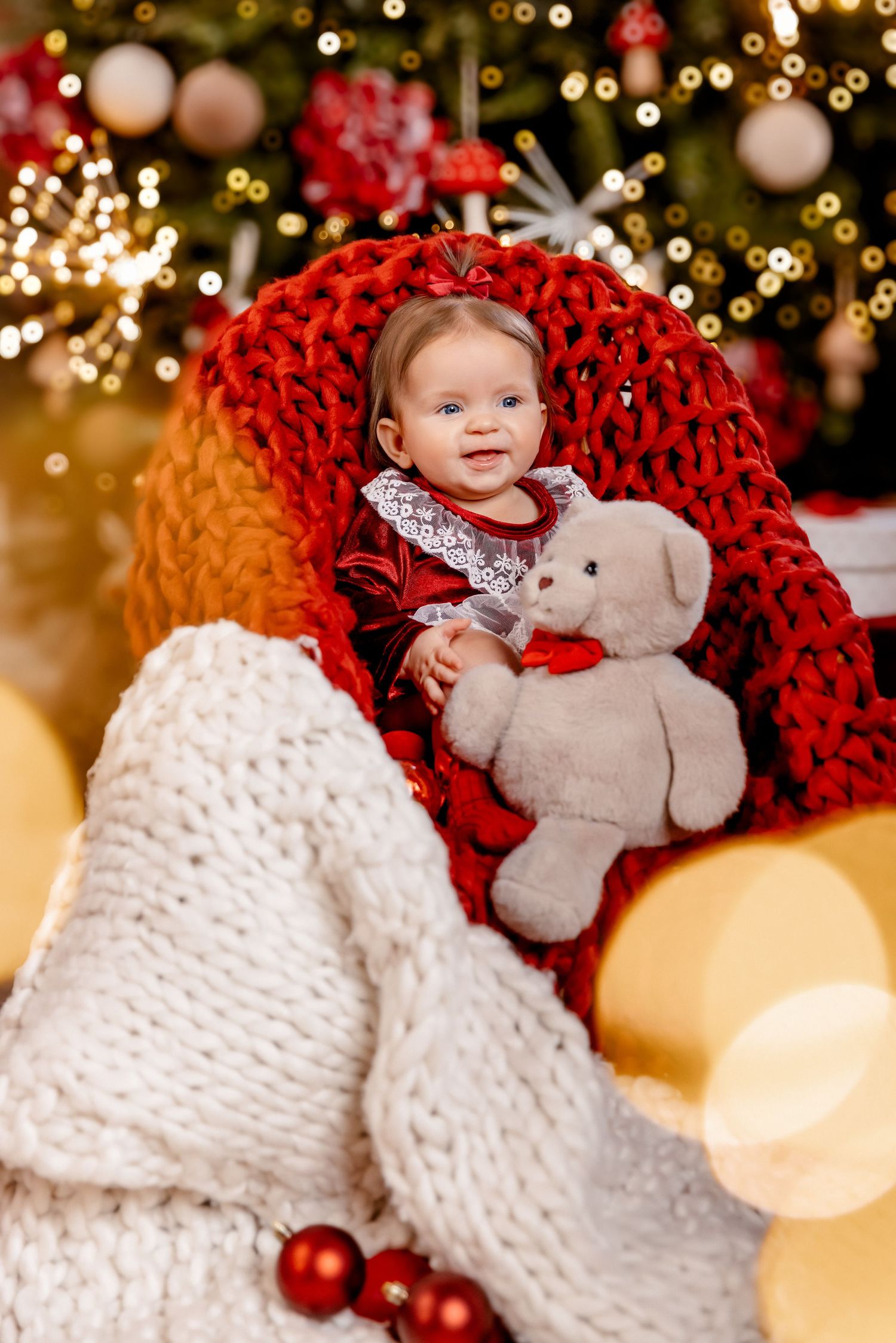 A smiling baby in a red dress holds a plush bear while sitting in a chunky red knit chair by a Christmas tree.